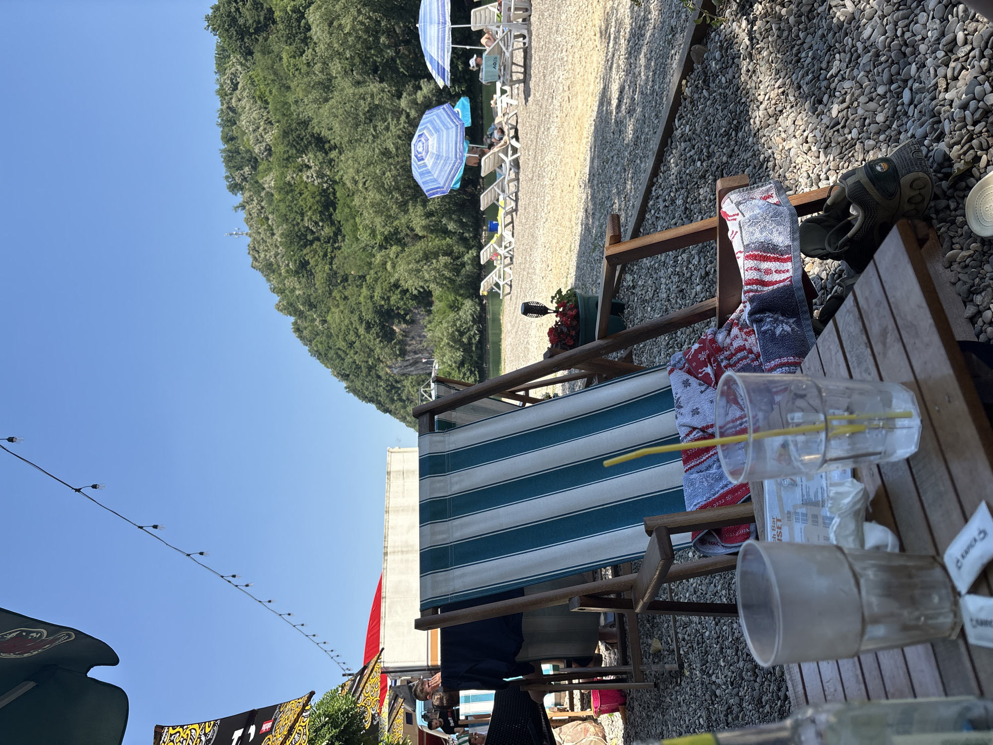This image depicts a sunny outdoor seating area at a beach bar, likely located in Zvornik, Bosnia and Herzegovina. In the foreground, a wooden table holds two empty drinking glasses, one with a yellow straw. A bottle is also partially visible on the left side of the table. A striped deck chair, featuring teal and white stripes, is prominent in the center, draped with a towel. Adjacent to the chair, a pair of hiking shoes rests on the gravelly ground. Behind the chair, several other deck chairs are set up on a sandy and pebbly beach, some occupied by individuals relaxing under blue and white umbrellas. The background reveals a lush, tree-covered hill sloping down towards a body of water. The sky is clear and blue. String lights are visible overhead, strung between unseen structures, suggesting an evening ambiance.