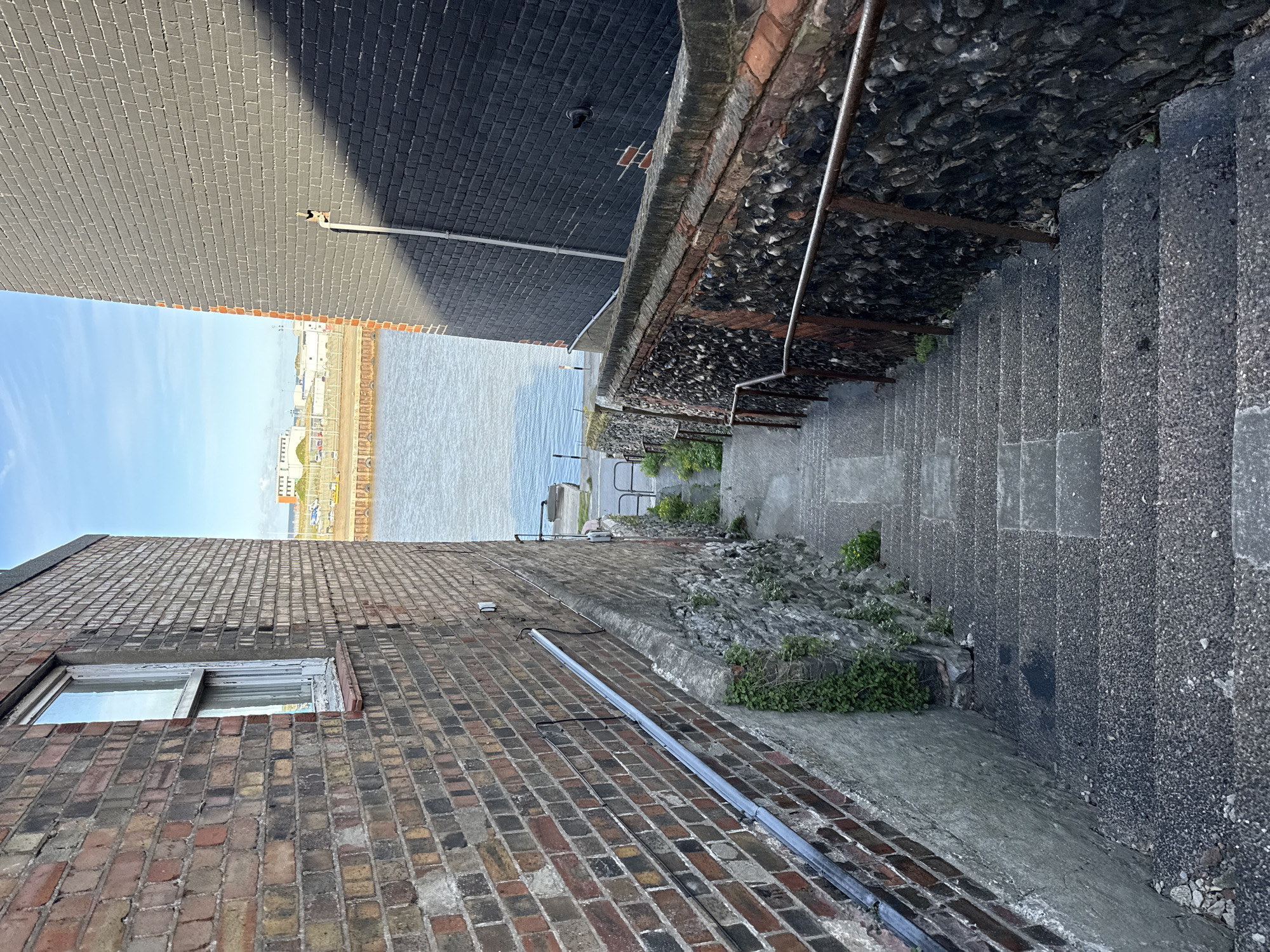 The image depicts an alleyway in Great Yarmouth, United Kingdom, leading down a steep set of stone stairs towards a body of water, likely the River Yare or the North Sea. The alley is flanked by brick buildings on both sides. The building on the left is constructed with a mix of red and brown bricks, and a partially visible window is present. A metal pipe runs along the building's facade. The building on the right is made of darker gray bricks. The stairs have a stone and concrete construction and are equipped with a metal handrail. Small patches of vegetation grow along the walls and between the stairs. At the bottom of the stairs, a partial view of a waterfront area with distant buildings and a clear blue sky is visible. The photo is taken from a slightly elevated perspective, looking down the alleyway towards the water.
