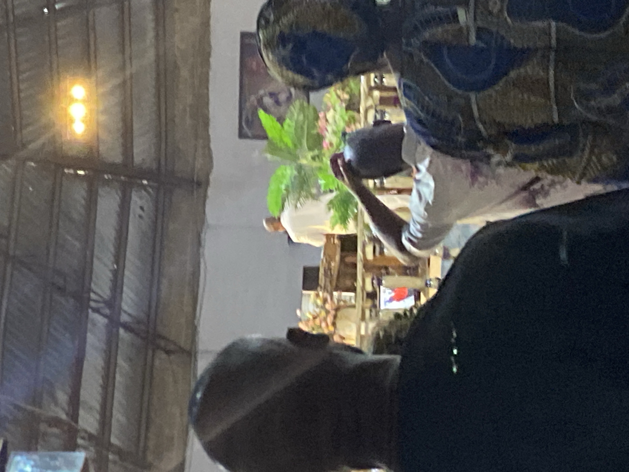 The image shows an interior scene at what appears to be a gathering, potentially in a large hall or structure, given the exposed rafters and metal beams visible overhead. A set of three vertically-aligned lights are visible in the background.
Several people are present, although their faces are mostly obscured or only partially visible. In the foreground, the back of a bald head and shoulder of a person wearing a dark shirt are noticeable. Towards the center, another person wearing a white shirt is seen holding a camera, possibly taking a picture of something beyond them. Above this person is another individual wearing a patterned shirt, but their details are not distinct. A framed image and some plants are also visible in the scene.
Given the location provided, Rukuba Road, Nigeria, the gathering might be related to a social, religious, or community event specific to the area. However, without more information, the exact nature of the event remains unclear.