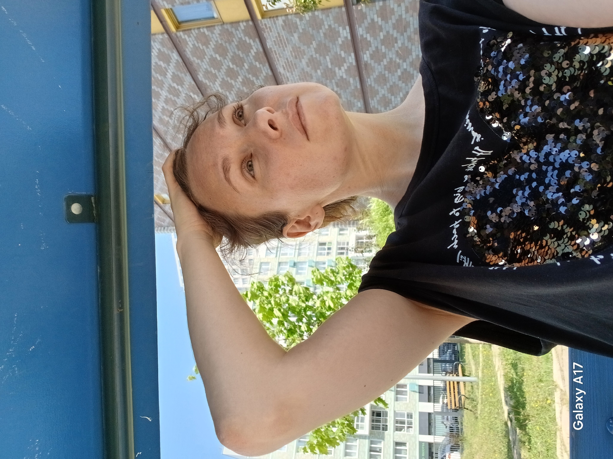 A medium-angle, horizontally-oriented outdoor shot shows a Caucasian woman with light brown hair and freckled skin standing under a blue-painted structure with a metal support bar. She is wearing a black t-shirt adorned with sequins. Her right arm is raised, with her hand touching her head. Behind her, there is a building with a geometric pattern, possibly a canopy or overhanging structure. The background features a green tree, a multi-story building, and a grassy area with a bench. The "Galaxy A17" watermark is visible in the lower right corner. Based on the urban elements such as buildings and infrastructure, and including the metadata "Yekaterinburg, Russia", the image was most likely taken in Yekaterinburg, Russia.