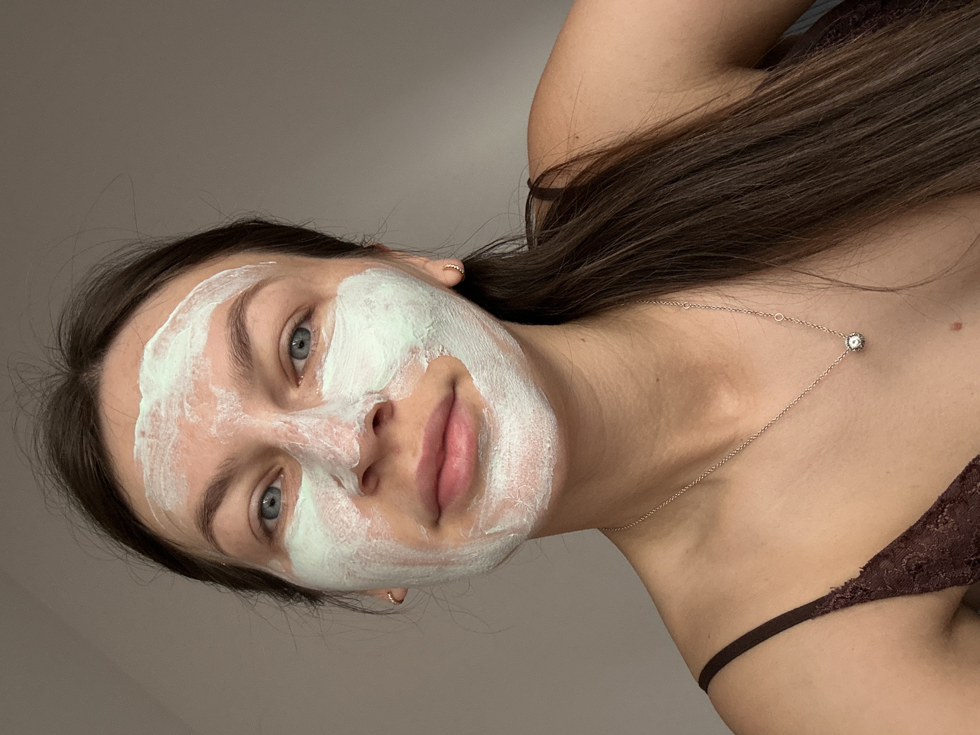A young woman with light skin is wearing a green-tinted white face mask. She has blue eyes and brown hair. She is wearing a gold necklace. The woman is wearing a brown, lacy top. The location is not discernible from the information provided.