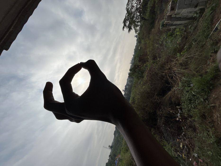 A human hand, silhouetted against an overcast sky, is positioned in the foreground, forming a circular gesture with the thumb and index finger. The sun's glare is visible through this opening. The remaining fingers are extended upwards. The scene, located in the city of Debasco, Nigeria, depicts a landscape beyond the hand comprising dense vegetation, including bushes and trees, receding into the background. A high-tension power line tower is visible on the distant left, above scattered residential rooftops nestled among hillsides. On the far right, a distinctive tree with a broad, flat canopy stands. In the lower right corner, a stack of concrete blocks is partially visible. The immediate ground below the hand consists of exposed soil and scattered debris.
