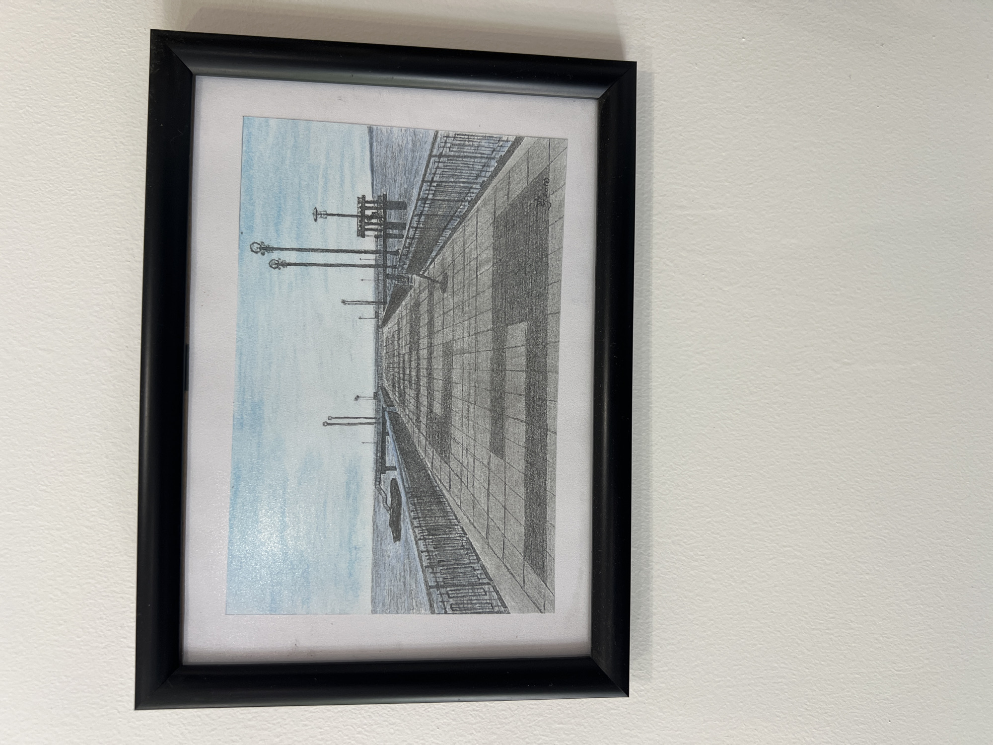 A framed, rectangular drawing hangs on a white wall. The drawing depicts an urban scene, oriented vertically. The frame is black. The drawing itself shows a wide, paved walkway or pier extending into a body of water, possibly a harbor or river. The pavement is rendered with repeating geometric patterns, suggesting tiles or blocks. Lamp posts are visible along the walkway. The body of water is colored with light blue. A boat is present on the water. The drawing has a perspective that makes the walkway seem to recede into the distance, creating a sense of depth. No people are visible in the scene. The location is unknown.