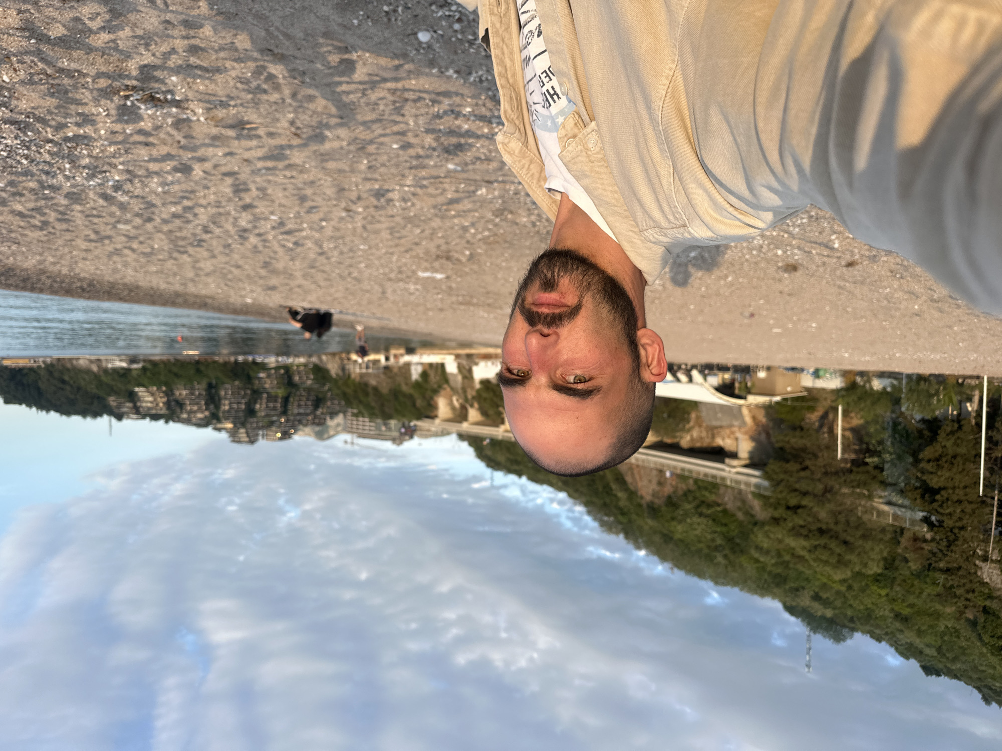 The image displays a close-up selfie of a man on a beach in Budva, Montenegro, taken during the golden hour. The man, with a short beard and mustache, and a neutral expression, looks directly at the camera. He is wearing a light beige jacket over a white t-shirt, on which fragmented text like "HYPER" and other letters are visible.
Behind the man is a wide, light brown sandy beach, extending towards a calm body of water. Small pebbles and shells are scattered across the sand. In the distance, near the water's edge, a few blurred figures are discernible, including one person in dark attire.
Beyond the water, the coastline features green, tree-covered hills dotted with numerous multi-story buildings, likely residential or hotel structures, characteristic of a coastal resort. The sky overhead is partly cloudy, illuminated by the warm, soft light of either sunrise or sunset. The scene conveys a peaceful moment by the sea.