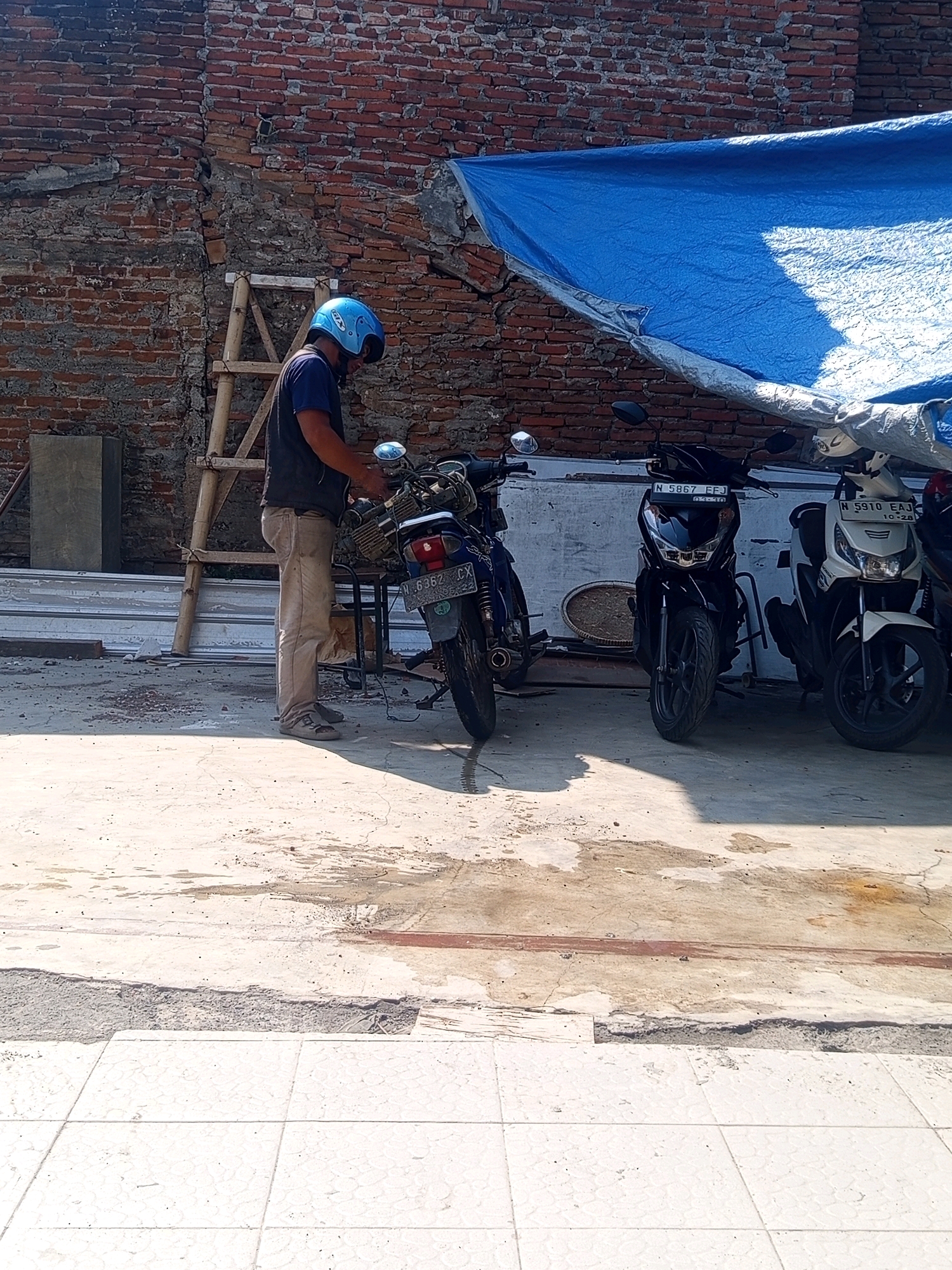 A man wearing a blue helmet and a dark vest over a blue shirt, stands next to a motorcycle. He is facing to his right and appears to be adjusting or working on the motorcycle. The motorcycle is dark colored with a license plate reading "N 6362 CX". Behind the man, a ladder leans against a brick wall that is partially covered by a blue tarp. To the right of the first motorcycle, two more motorcycles are parked, one black with license plate "N 5867 FFJ", and one white with license plate "N 5910 EAJ". The ground is paved with concrete and some areas are wet, with puddles and stains. The overall scene suggests an outdoor area, possibly a courtyard or alleyway, in a city environment. The brick wall and the style of the motorcycles suggest a location in Malang, Indonesia.