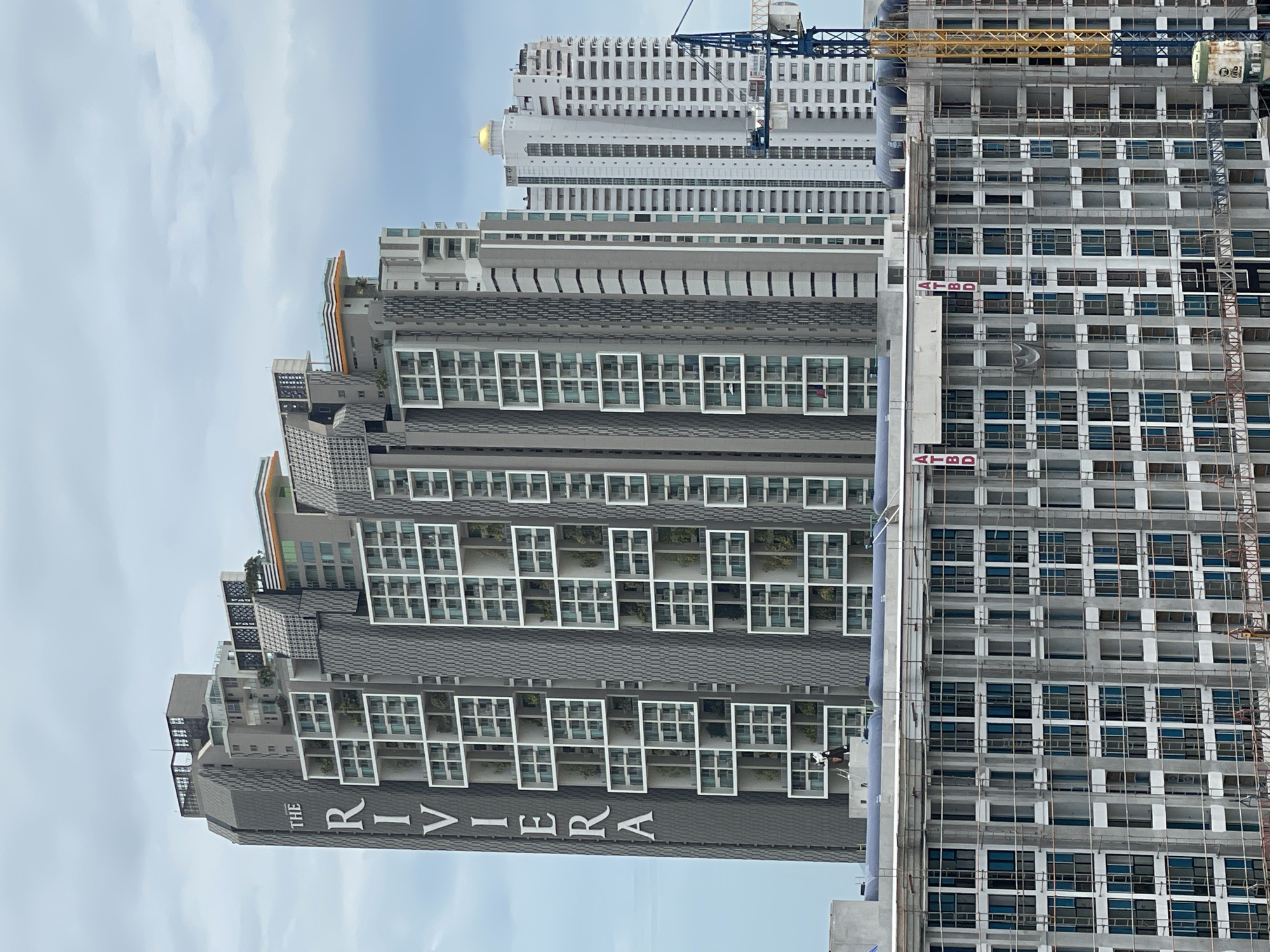 This image shows a construction site in Pattaya City, Thailand, dominated by a large, modern skyscraper complex. The main building, "THE RIVIERA," features a stepped design with numerous balconies and windows, some of which contain greenery. To the right, other high-rise buildings are visible, including one with a golden dome. In the foreground, a multi-story building is under construction, characterized by scaffolding and visible interior structures. A blue construction crane is positioned on the right side of the image, adjacent to the partially built structure. The sky is overcast with clouds.