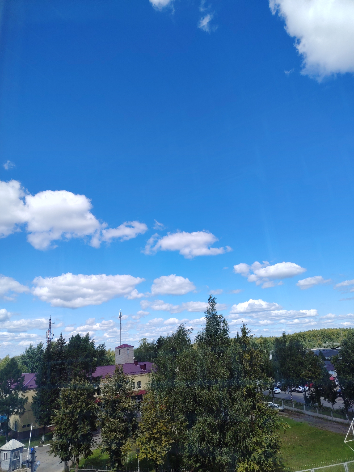 The image shows a clear blue sky with scattered white clouds above a landscape that includes buildings, trees, and a parking lot. In the foreground, there is a small, gray guard booth with a white railing and a gate. Behind the booth, a light-colored building with a burgundy roof is visible, along with several tall evergreen trees. Further back, another building with a burgundy roof and a tower is present, flanked by more trees. A parking lot with several white and red cars is situated to the right of the buildings. The background consists of a dense forest. The scene appears to be a sunny day in a suburban or semi-rural area.