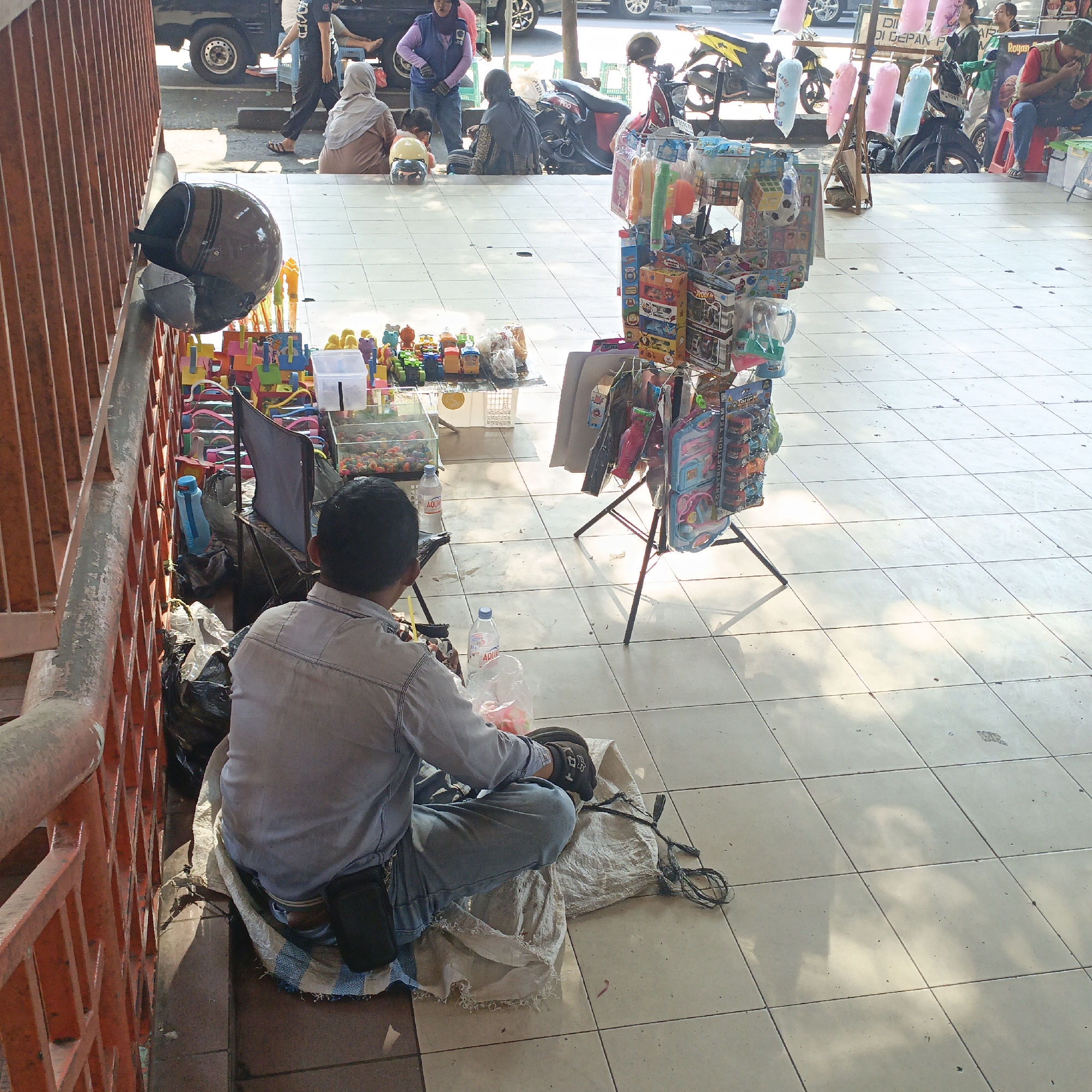 This image depicts a street scene in Salatiga, Indonesia, likely a sidewalk adjacent to a road. In the foreground, a man is seated on a makeshift cushion on the tiled ground, facing away from the camera. He is wearing a light blue long-sleeved shirt and blue jeans. His right hand, wearing a dark glove, is engaged with something on his lap. A black phone holster is attached to his belt. To his left, a black helmet rests on a red railing. In front of him are several bottles of water and plastic bags containing items, possibly candy or small toys.
Behind the man, a display rack holds various toys, including toy cars and dolls, attracting potential customers. Further back, several people are visible, some seated on the ground and others standing. One woman, wearing a hijab and a purple top, appears to be interacting with a child. Another person in a hijab is seated on the ground. A motorcycle is parked nearby. In the background, more people and a parked car suggest a busy urban environment. The ground is covered with light-colored square tiles, and sunlight casts shadows, indicating it is daytime. The overall scene suggests a bustling marketplace or roadside vendor area.