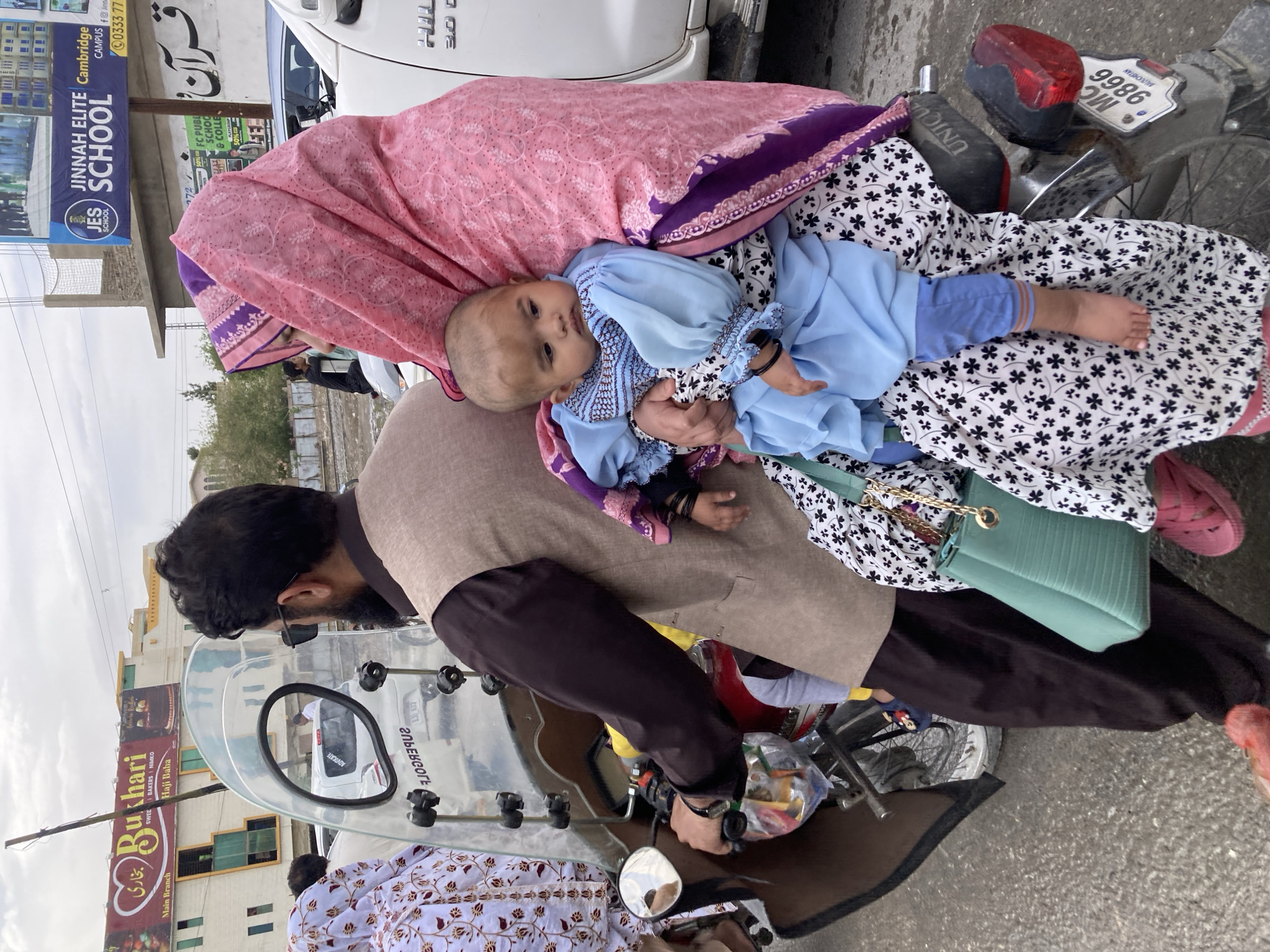 The image shows a family on a motorcycle in an urban street setting, identified as Quetta City Tehsil, Pakistan, under an overcast sky during daytime. A bearded man, wearing a brown shirt and black sleeves, is seated on the motorcycle, holding the handlebars. Behind him, a woman, mostly covered by a pink patterned headscarf and a black and white patterned dress, is holding a baby. The baby, dressed in a light blue outfit, looks towards the camera. A light green handbag with a gold chain rests on the woman's lap. Another person in a white patterned outfit is partially visible on the motorcycle.
The motorcycle features a clear windshield, a rearview mirror (reflecting the word "ADVOCATE"), and a rear number plate displaying "MC 9866 BALOUCHISTAN," confirming the location. A white HILUX 3.0 vehicle is parked behind the motorcycle. The street is paved with asphalt, and various commercial buildings line the roadside.
Multiple signboards are visible, including "JINNAH ELITE SCHOOL Cambridge CAMPUS JES SCHOOL," "FC PUBLIC SCHOOL & COLLEGE 50%," and "Bukhari SWEET BAKERS NIMKO Haji Baba Main Branch." A partial phone number "0333 77" is also seen, along with the word "قرآن" (Quran) in Arabic/Urdu script. The scene depicts a common mode of family transport in the region.