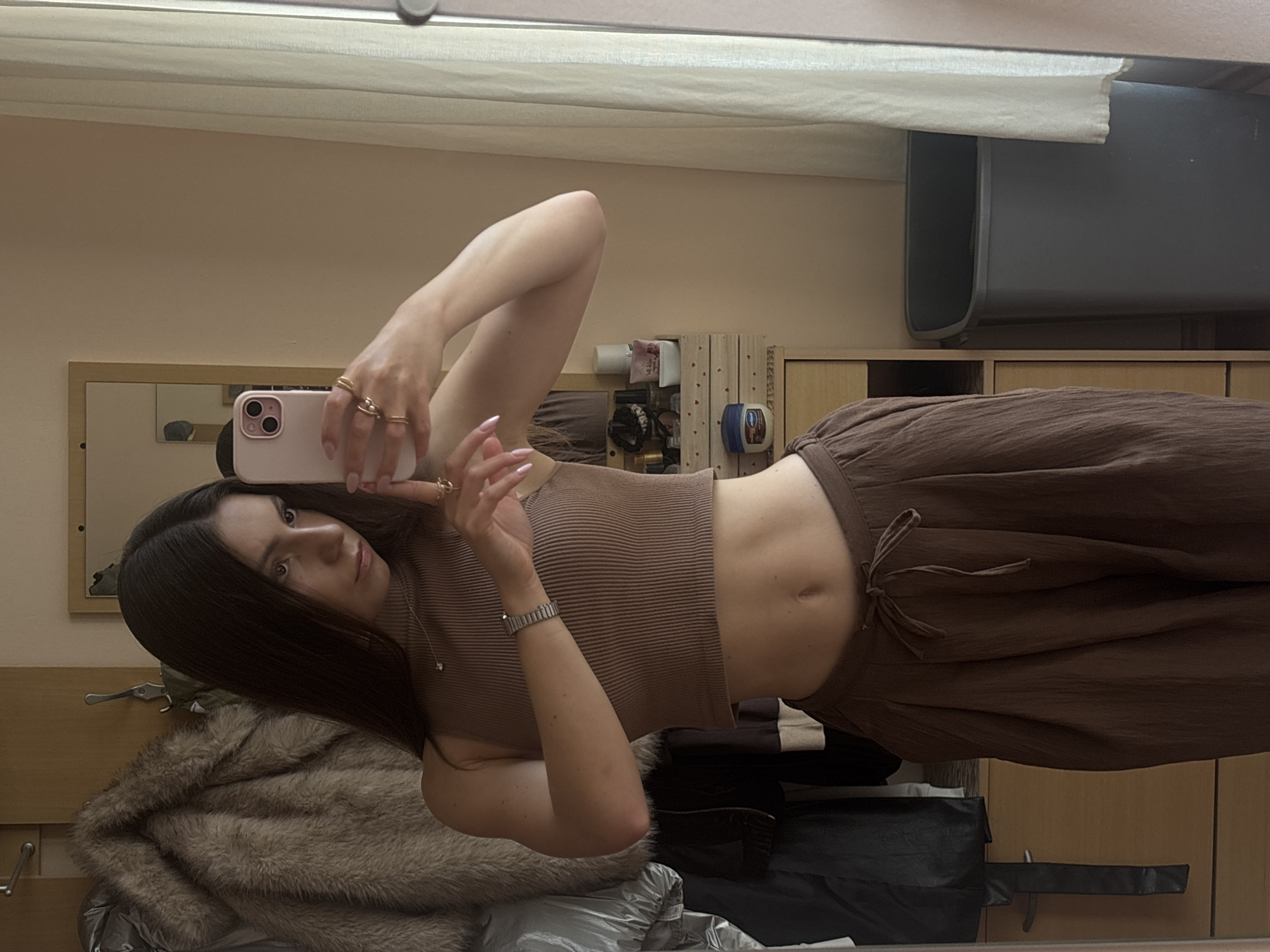 A young woman with long, dark hair is taking a mirror selfie in what appears to be a bedroom. She is wearing a brown ribbed tank top that exposes her midriff and loose, brown, textured pants tied at the waist with a drawstring. She is wearing several rings on her fingers and a watch on her left wrist. Her fingernails are painted a light pink. Her expression is neutral, and she is looking directly at her reflection in the mirror.
The background shows a wooden dresser or wardrobe, a shelf with various items including toiletries and what looks like a small wooden organizer, and a large grey bin. A fluffy fur coat is draped over a coat rack on the left side of the image. A mirror is mounted on the wall, reflecting the woman and the room. The lighting suggests it could be daytime, though no windows are clearly visible. The overall impression is of a casual, indoor setting.