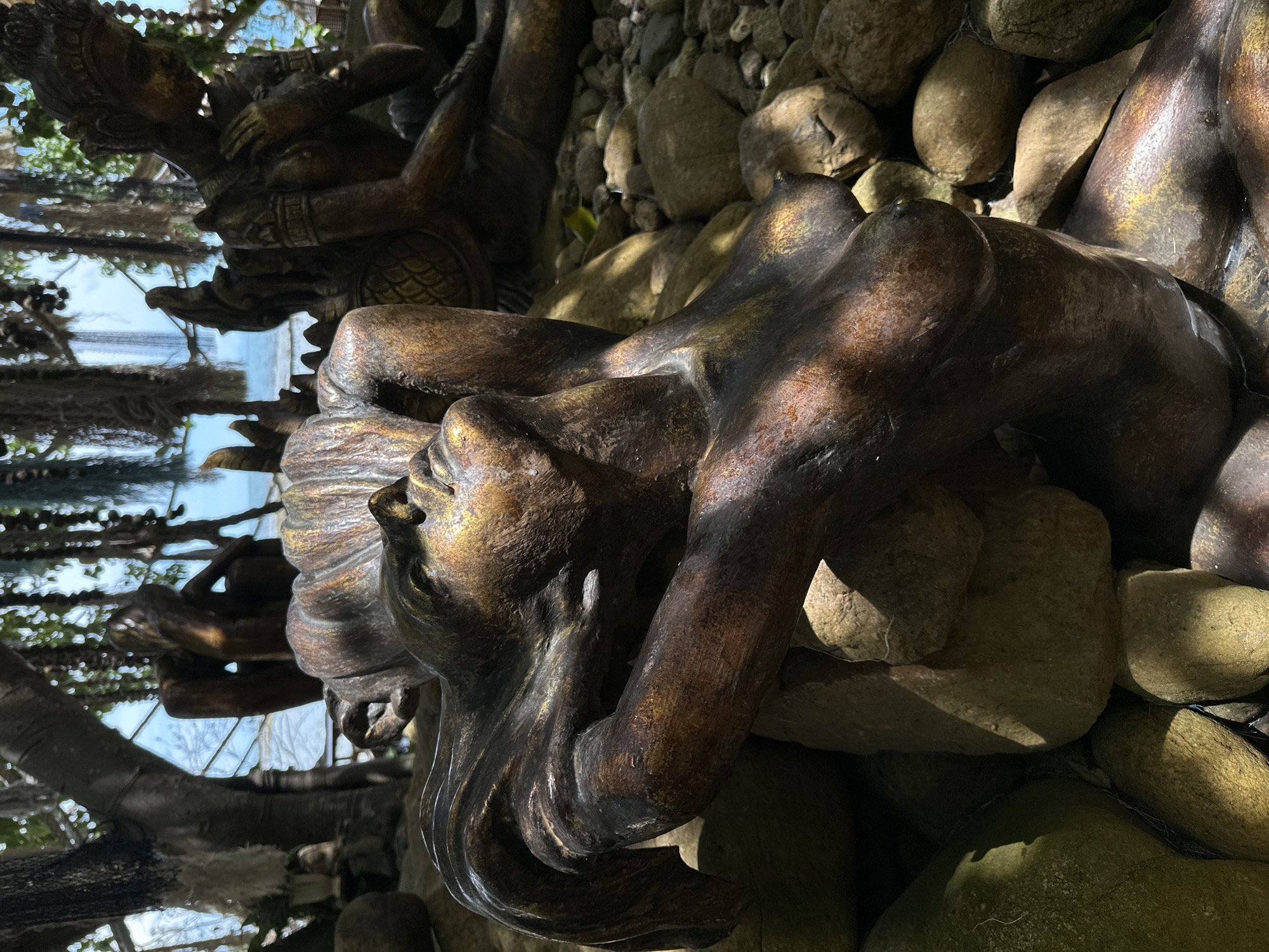 The image displays a close-up of a bronze sculpture of a reclining female figure, possibly a mermaid or siren, resting on a bed of river stones. The sculpture has a weathered, metallic finish with areas of gold patina. The figure's head is tilted back, with her arms supporting her head, and her gaze directed upwards. The detailed rendering of her form is evident, including her torso and flowing hair. In the background, other sculptures are partially visible, along with lush green foliage and a glimpse of the ocean under a bright sky. The setting appears to be an outdoor garden or display area, possibly a tourist attraction. The overall impression is one of artistic display and natural beauty.