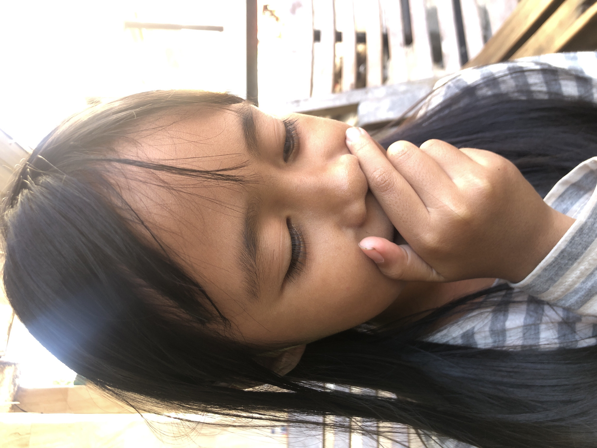 A young girl with long, dark hair is looking down and covering her nose and mouth with her right hand, her eyes closed. She is wearing a grey and white striped shirt. The background appears to be outdoors, with blurred wooden structures and bright sunlight filtering in, suggesting a location in Surabaya, Indonesia.
