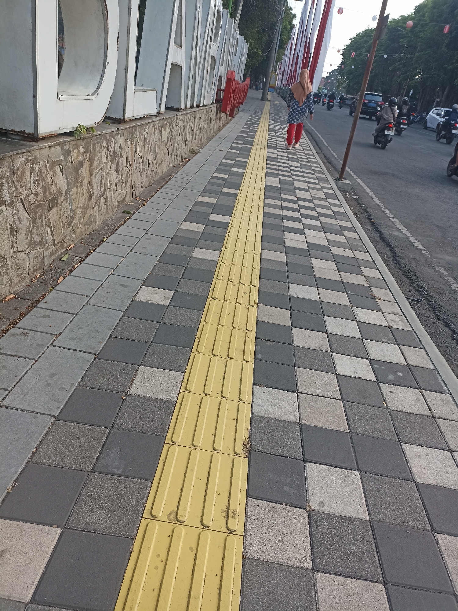 A pedestrian sidewalk with tactile paving is depicted in the foreground, situated next to a street with light traffic, including several motorcycles and a car. The sidewalk is paved with alternating grey and white square tiles, with a bright yellow tactile paving strip running along its edge. A person wearing a headscarf, a patterned top, and red trousers walks away from the camera on the sidewalk. Behind the sidewalk, a low stone wall runs horizontally, and large white letters spelling out a word are visible, with the partial word "DIAN" clearly decipherable. Red and white banners are strung across the street in the background. The scene appears to be in a moderately urban environment, possibly a public walkway or promenade in Kelurahan Kepatihan, Indonesia, given the context of the street traffic and pedestrian infrastructure. The lighting suggests it is daytime, with some shadows present.
