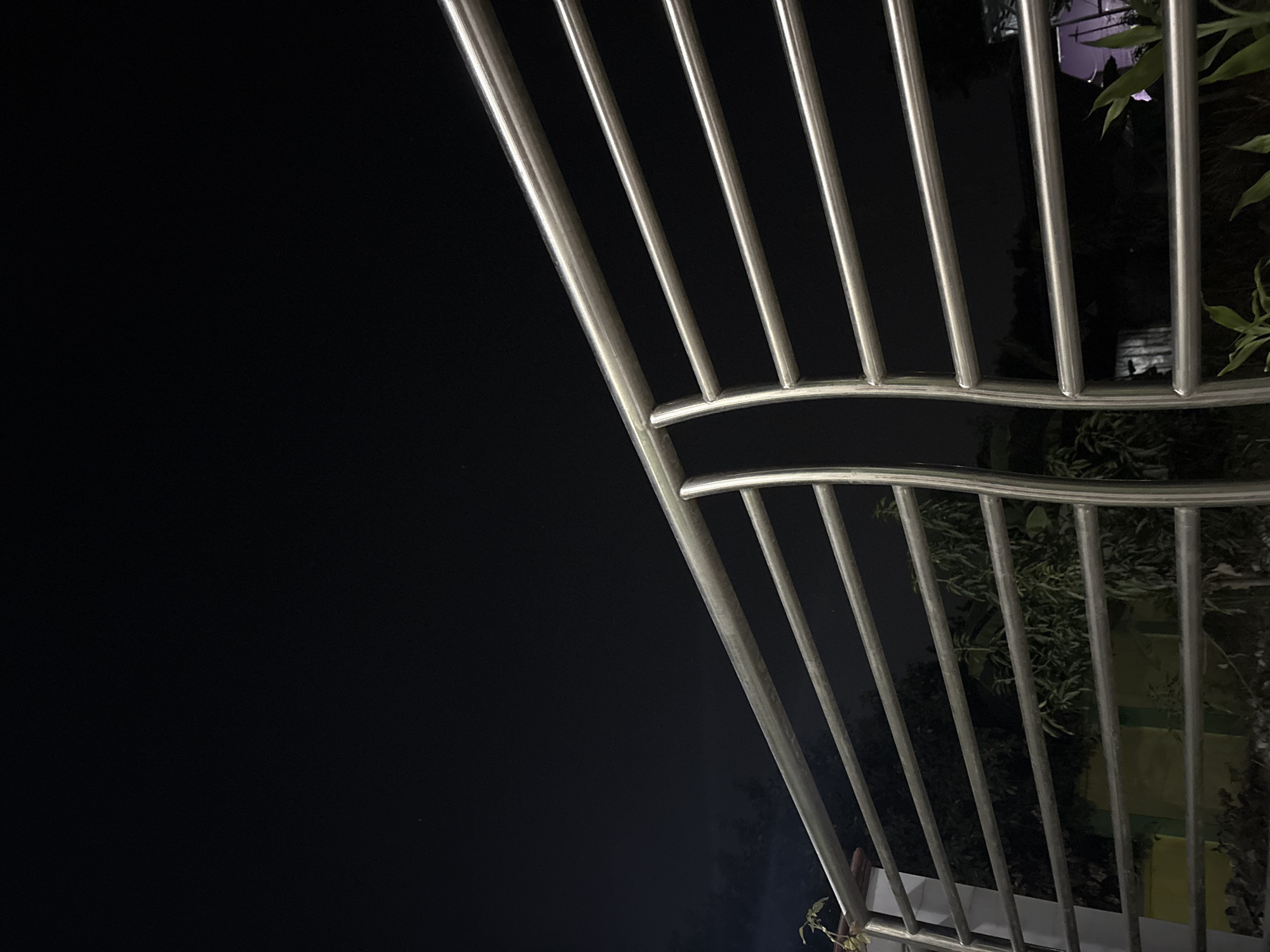 A nighttime exterior shot shows a metal railing in the foreground, with the dark sky and some faint greenery visible behind it. The railing is composed of horizontal bars and has a curved support in the center. The location is the city of Gempol, Indonesia. The image is lit by artificial light, creating a stark contrast between the illuminated railing and the dark surroundings. There are no people or animals visible in the image.