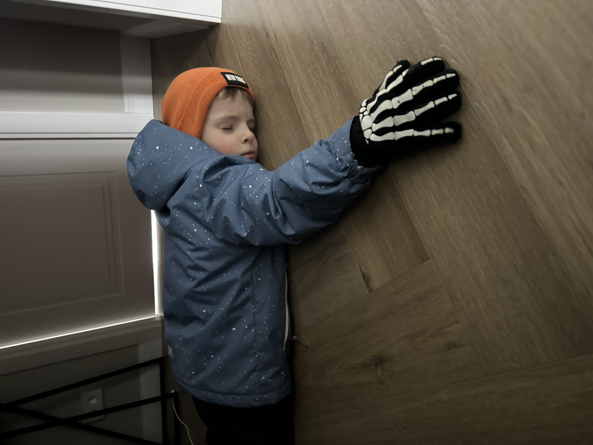A child wearing a blue winter jacket with white specks, an orange knit cap with a dark rectangular patch, and a black glove with white skeleton bone design lies on a wood-look floor. The child's left arm, encased in the skeletal glove, presses against the floor. The child's eyes are closed. The location is inside a building in the city of Chaikovsky, Russia. A white door with panels is visible in the background.