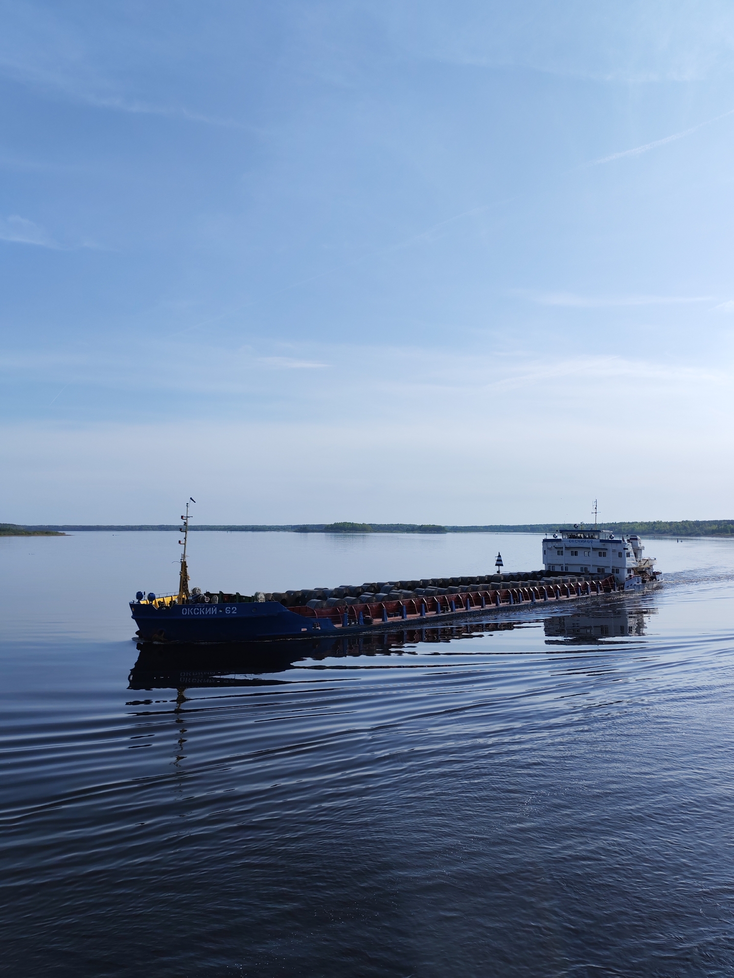 The image shows a cargo ship, named "ОКСКИЙ - 62", moving on a calm body of water. The ship appears to be carrying cylindrical cargo. The sky is clear with a few wisps of clouds. The shoreline is visible in the background with green vegetation. The location is likely near Shelomovo, Russia.