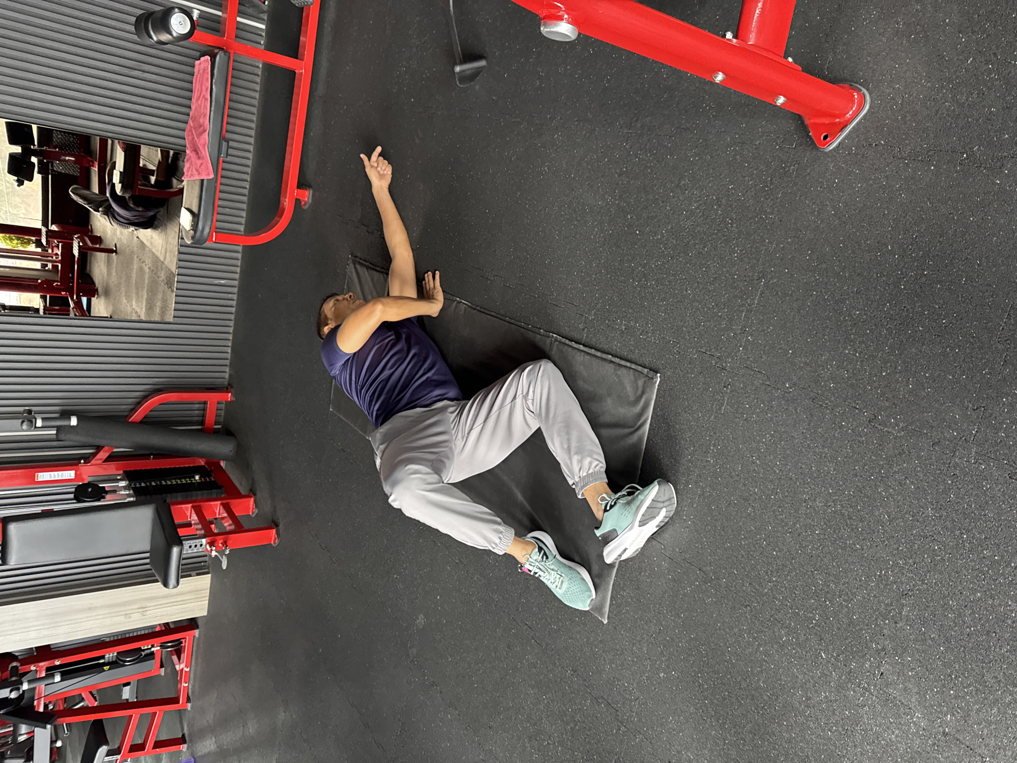 A man lies on his back on a black mat on a gym floor, performing a stretch. He is wearing a dark blue t-shirt and light grey sweatpants. His legs are bent, and he is reaching his right arm out to the side, with his index finger extended. The gym features red weight machines and equipment, with a grey, ribbed wall in the background. A mirror reflects more of the gym equipment. The flooring is a dark, textured rubber. The image appears to have been taken in Kolkata, India, as indicated by the context provided.
