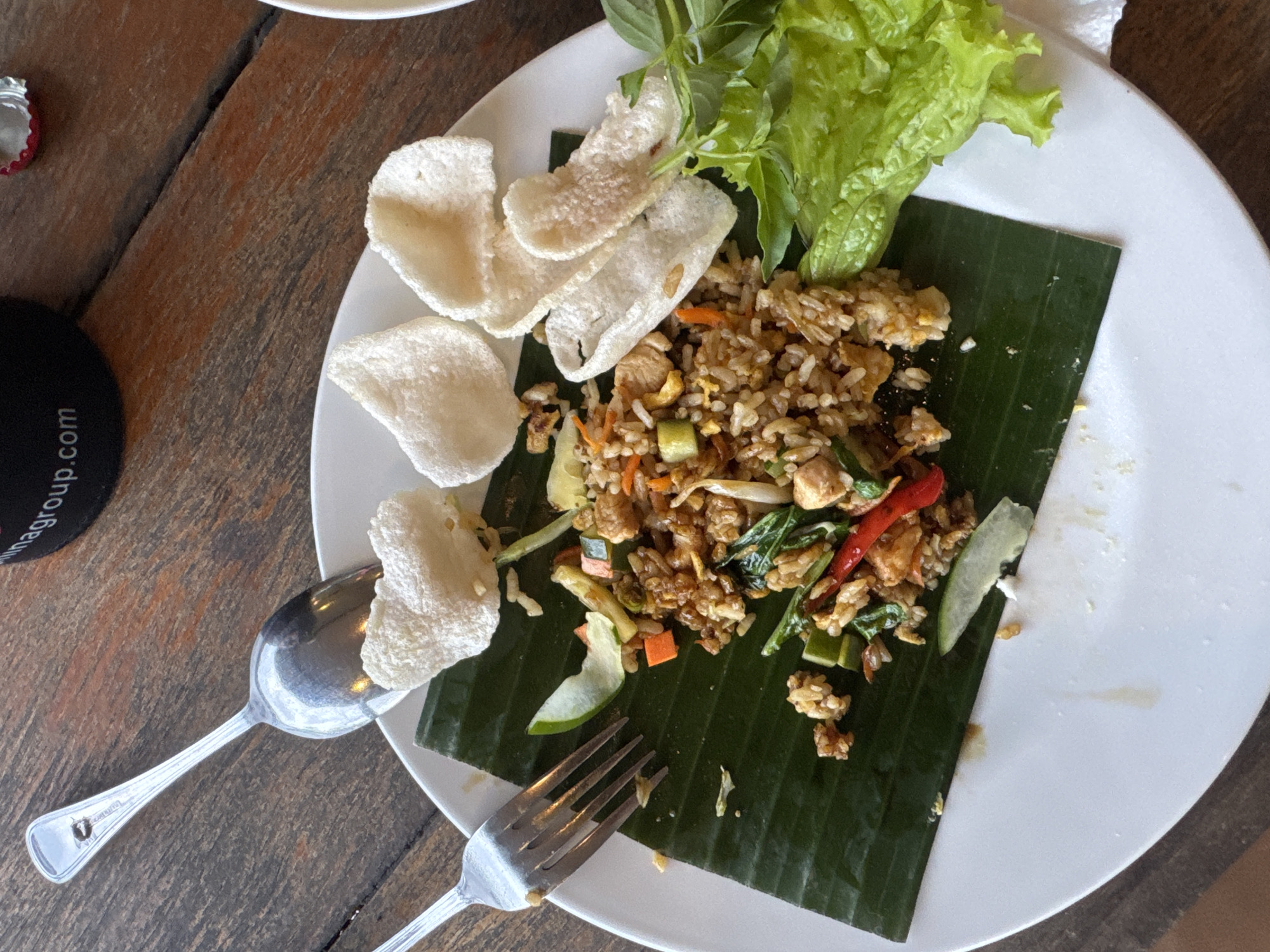 The image shows a plate of nasi goreng, an Indonesian fried rice dish, served on a large white plate atop a wooden table. The meal is presented on a section of banana leaf, adorned with lettuce, crackers, and basil. A fork and spoon rest beside the plate. A black coaster is also partially visible on the table. Based on the metadata, the image was taken in Baturiti, Indonesia.