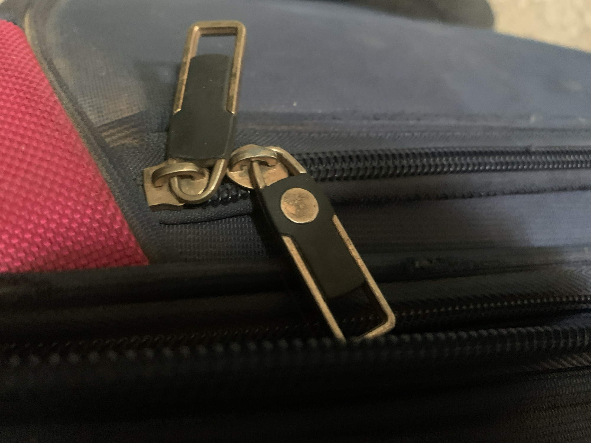 A close-up, high-angle shot shows two gold and black zipper pulls on a navy blue suitcase. The zipper pull on the left is a rectangular loop with a black rectangular tab. The zipper pull on the right is a smaller, elongated rectangular tab with a circular detail at its base. The suitcase fabric appears textured, and a sliver of bright pink fabric is visible on the left side of the frame. The image is focused on the zippers and the immediate surrounding fabric, with the background slightly blurred. The scene is indoors, on a suitcase. There is no indication of its location in Jalingo, Nigeria.