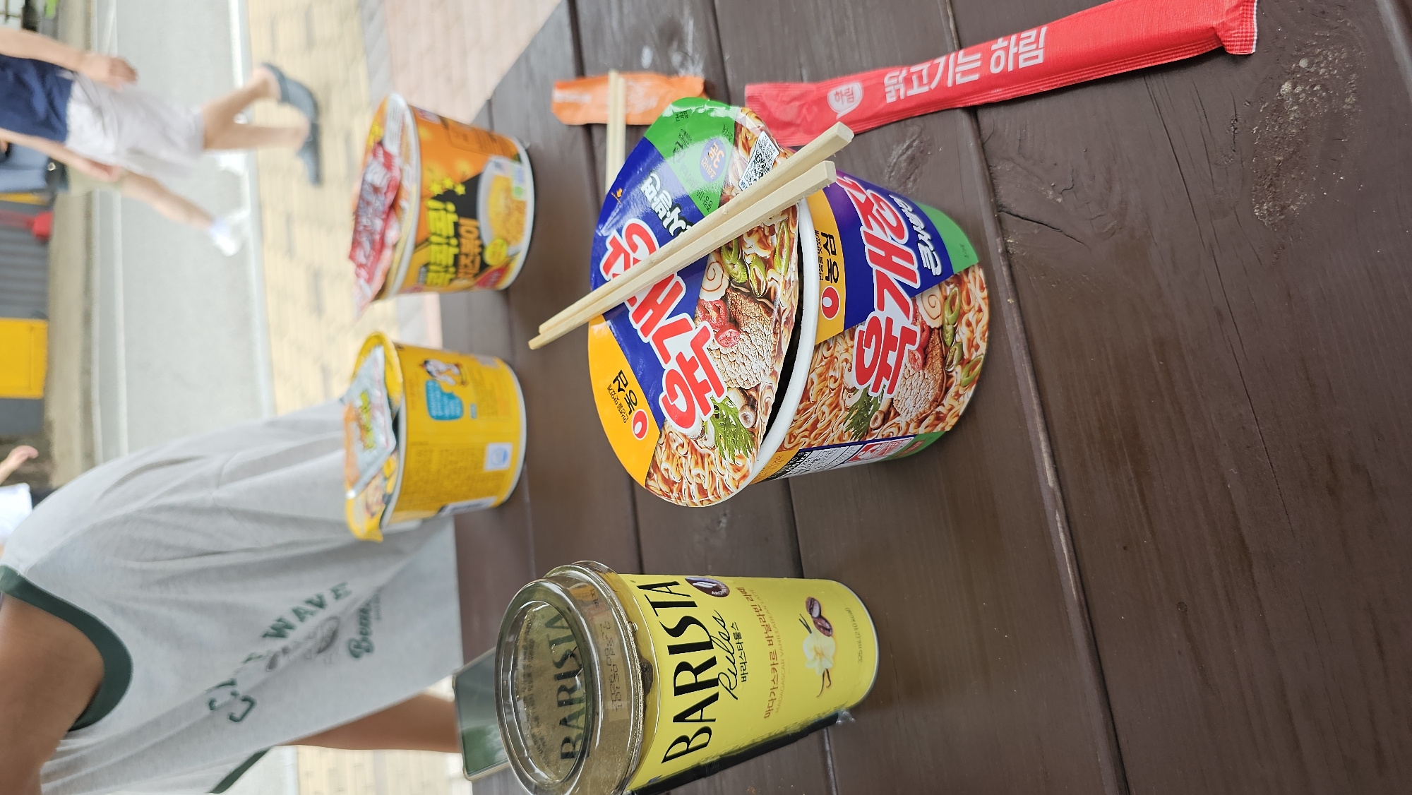 At a low-angle, eye-level shot, three cups of instant ramen and a cup of iced coffee are arranged on a wooden picnic table. The ramen cups are from the brand "Yukgaejang," featuring red packaging with images of noodles, meat, and vegetables. One cup of ramen is open, with chopsticks resting on top. The iced coffee, a "Barista Rules" vanilla latte, is in a transparent plastic cup. In the background, a person wearing a white t-shirt with green trim is partially visible, along with other blurred figures and a yellow structure. The setting appears to be an outdoor public space, possibly near a park or recreational area in Asan-si, South Korea.