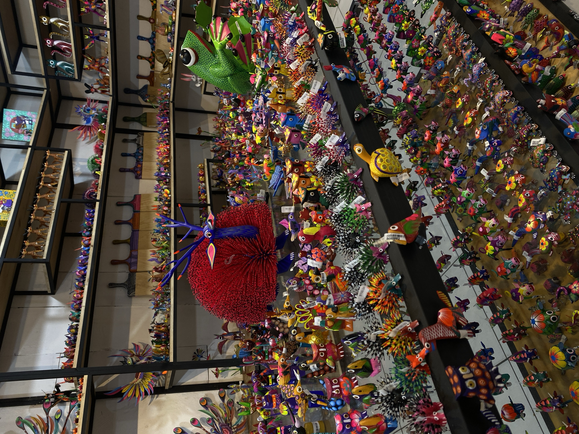 The image depicts a vibrant display of colorful alebrijes (Mexican folk art sculptures of fantastical creatures) in Oaxaca City, Mexico. The alebrijes, primarily animals and mythical beings, are densely packed on shelves and wall-mounted displays. They are varied in size, from small figurines to larger, more elaborate pieces. The shelves are constructed of dark metal frames and wood panels, creating a grid-like structure for displaying the alebrijes. Price tags are visible on many of the items, indicating they are for sale. The scene appears to be a shop interior, showcasing the artistic craftsmanship and cultural heritage of the region.