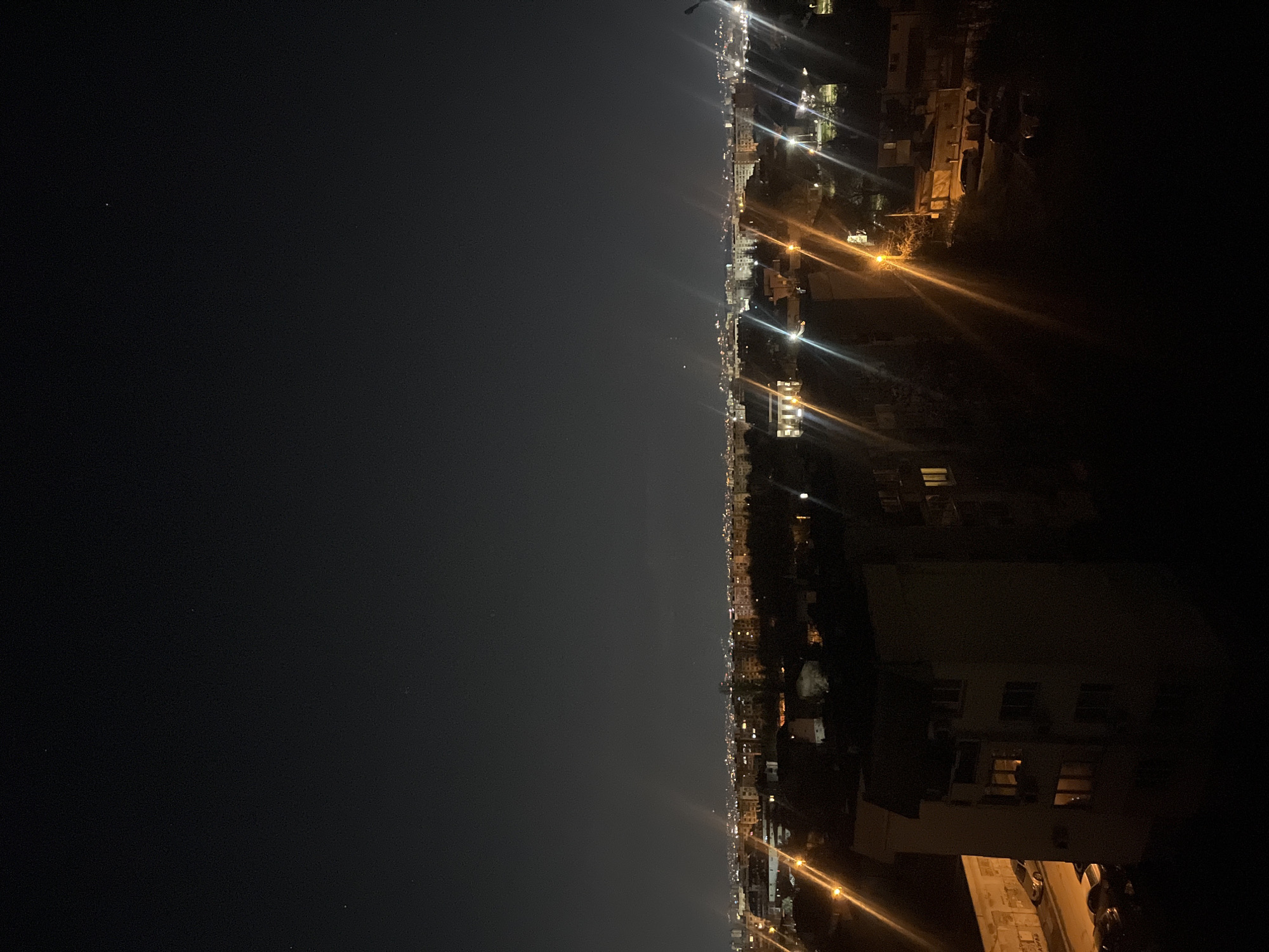 The image shows a night view of Sofia, Bulgaria from a high vantage point. On the right side of the image is a neighborhood with buildings and lit streetlights. The left side is a dark sky, suggesting it is late at night. There are no people visible. The camera angle is unusual, rotated 90 degrees counterclockwise. The environment is urban with a dark nighttime setting.