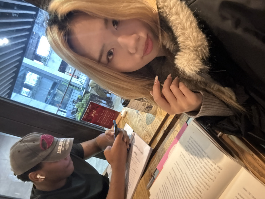An indoor scene depicts two individuals seated at a wooden table within what appears to be a cafe or restaurant in Yuzhong District, China. In the foreground, a female of East Asian descent with long blonde hair and dark roots, wearing a black jacket with a prominent faux fur collar and a grey ribbed sweater, directly faces the camera, holding her right hand near her chest. Her nails are manicured with a subtle design. To her left and slightly behind, a male of African descent, wearing a dark t-shirt and a grey baseball cap worn backward (featuring a pink design and "RADBLOOD" text), is engrossed in a black smartphone held in both hands. A white earbud is visible in his left ear.
On the wooden table between them are several open books or binders displaying Chinese text, with one featuring a pink divider. A white AirPod charging case and a disposable dark-colored coffee cup with white branding are also on the table. Through a large window in the background, an outdoor or semi-outdoor area is visible, featuring green foliage, architectural elements, and a red vertical sign with Chinese characters and the English text "Happy New Year 2024," along with what appear to be menu items or prices. The lighting suggests daytime, with a soft overcast sky reflected in the distant building.