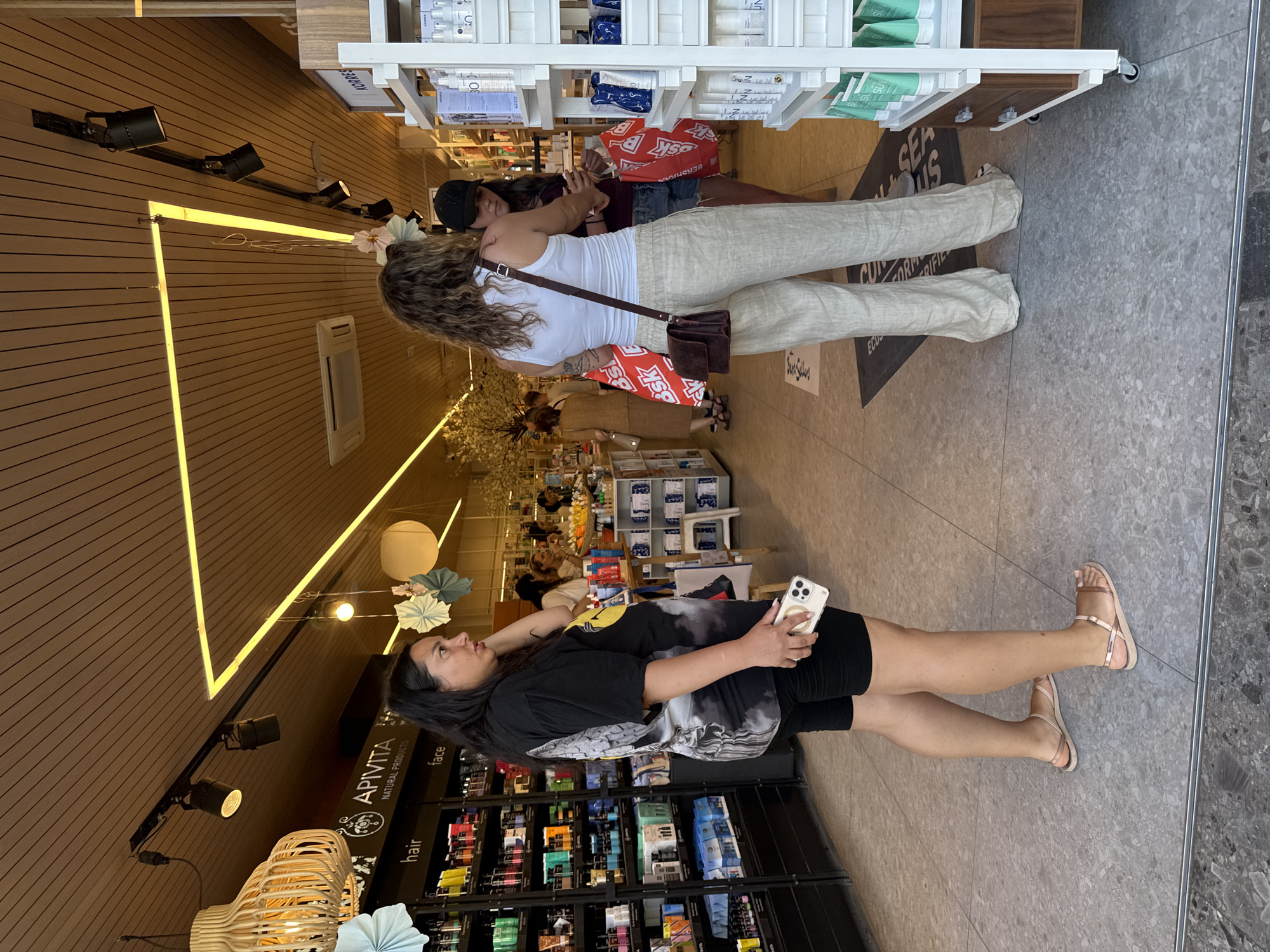 The image captures an indoor scene of a retail establishment in Chania, Greece, likely a pharmacy or boutique selling beauty and personal care products. The store features wooden slat walls and ceiling, illuminated by a modern, angular yellow light fixture and several track lights. Shelving units stocked with various products are visible, including a prominent display of "APVITA NATURAL PRODUCTS" on the left.
In the foreground, a woman with dark hair, wearing a black t-shirt and black shorts, stands facing right. She is holding a smartphone in her right hand and appears to be engaged with something on the device. To her right, another woman with wavy blonde hair, dressed in a white tank top and beige linen pants, is standing and looking towards a third woman. The second woman has a brown crossbody bag and is holding two red shopping bags with white lettering. The third woman, wearing a black baseball cap, is looking down at something in her hands, possibly also a phone or product.
The flooring is a gray, speckled tile. Decorative paper fans are suspended from the ceiling, adding a festive touch. The overall atmosphere suggests a bright and well-lit commercial space.