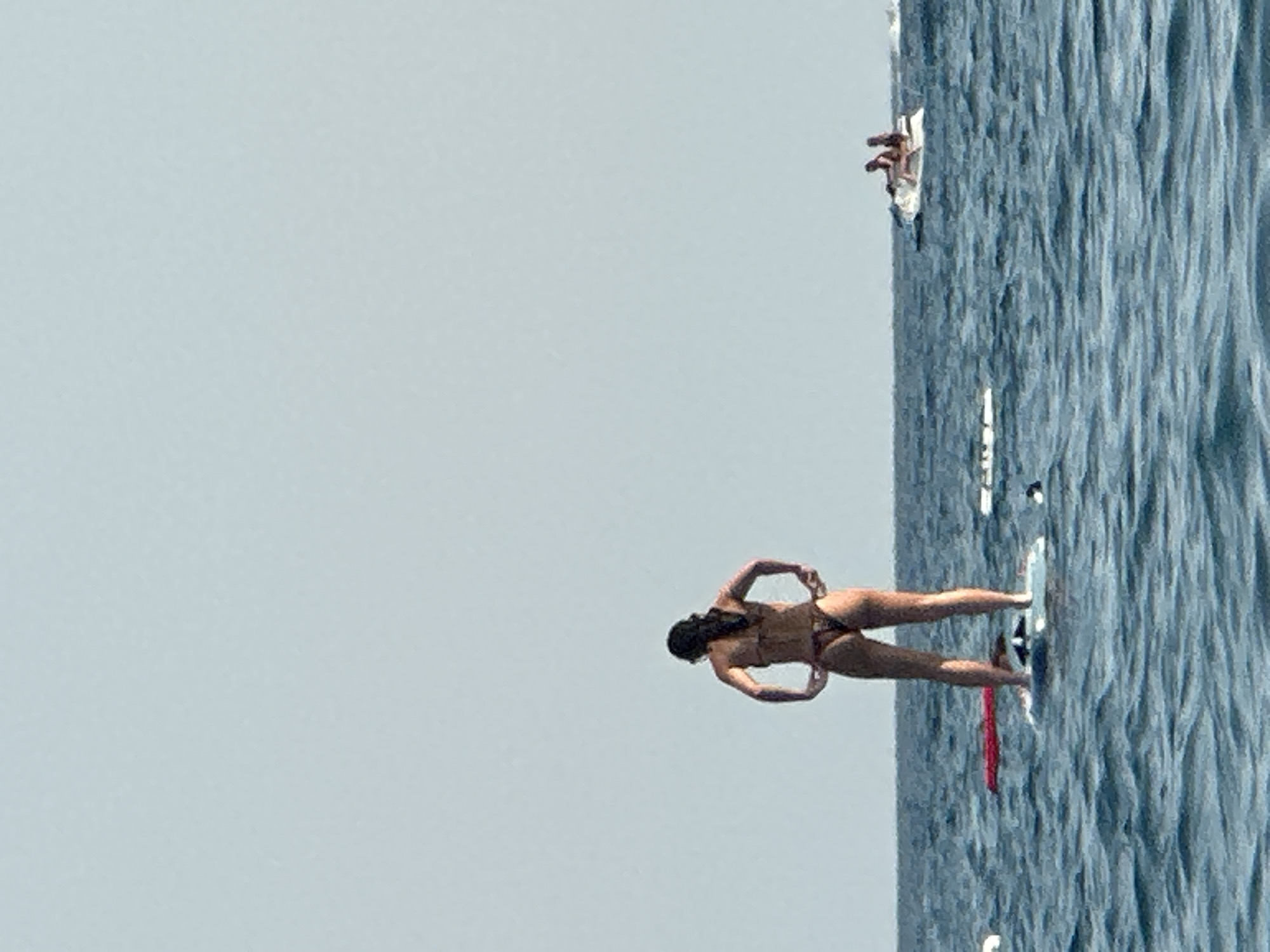 In Boreti, Montenegro, on a body of water with choppy blue waves under a clear, light blue sky, a woman stands on a paddleboard. She is viewed from behind, wearing a dark-colored bikini. Her hands are placed on her hips, and her hair is pulled back. To the right, further out on the water, two people ride on a jet ski, and to the left, another paddleboard is visible.