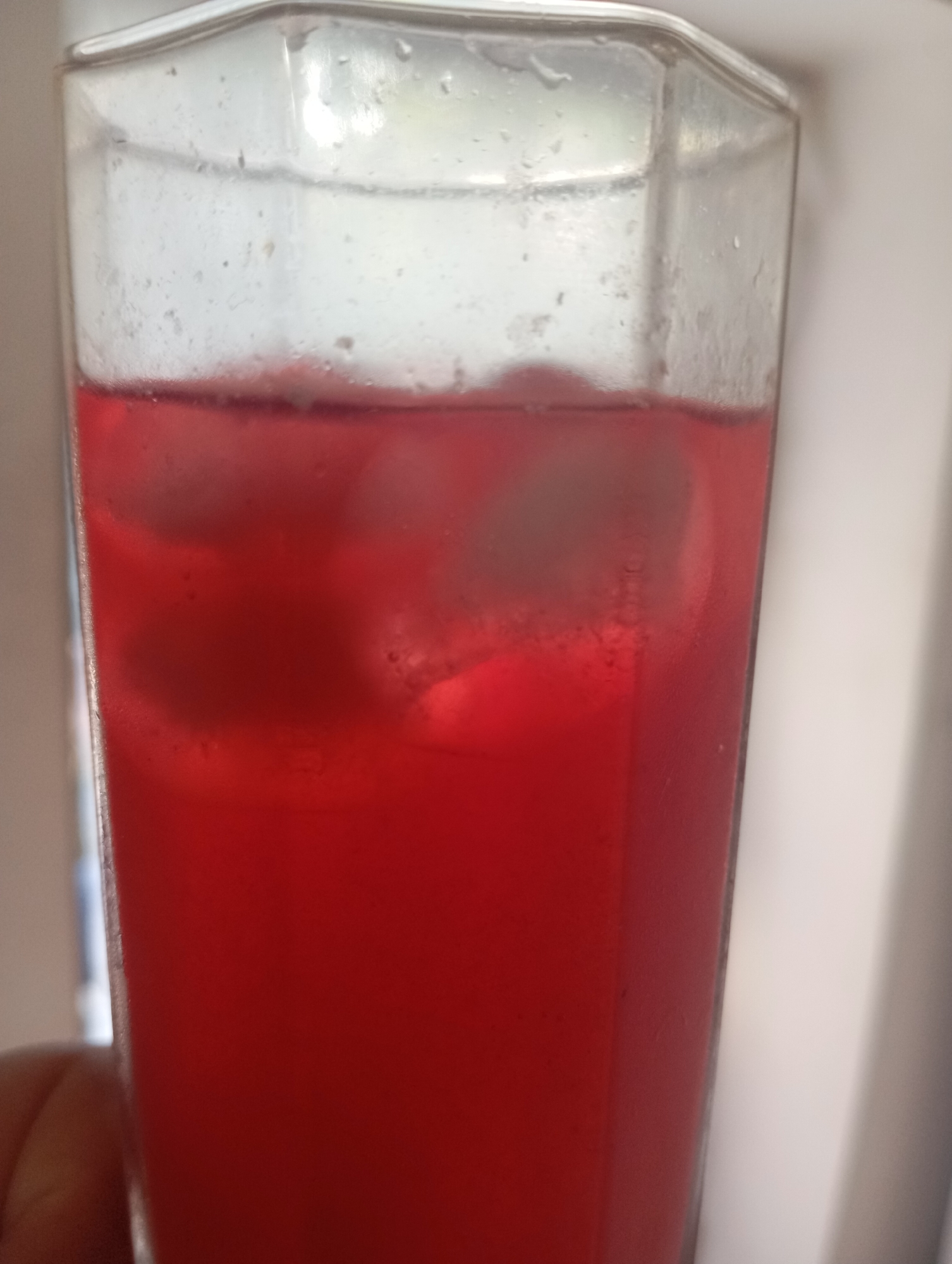 A close-up shot shows a glass of red liquid with ice cubes. The glass is tall and has a faceted design. The liquid is a vibrant red color, and several ice cubes are visible within it. The glass appears to be slightly frosted on the outside. The background is blurred and shows a light-colored wall or curtain, suggesting an indoor setting. A person's thumb is visible in the lower left corner, holding the glass. The image does not provide any specific details about the location in Almaty, Kazakhstan.