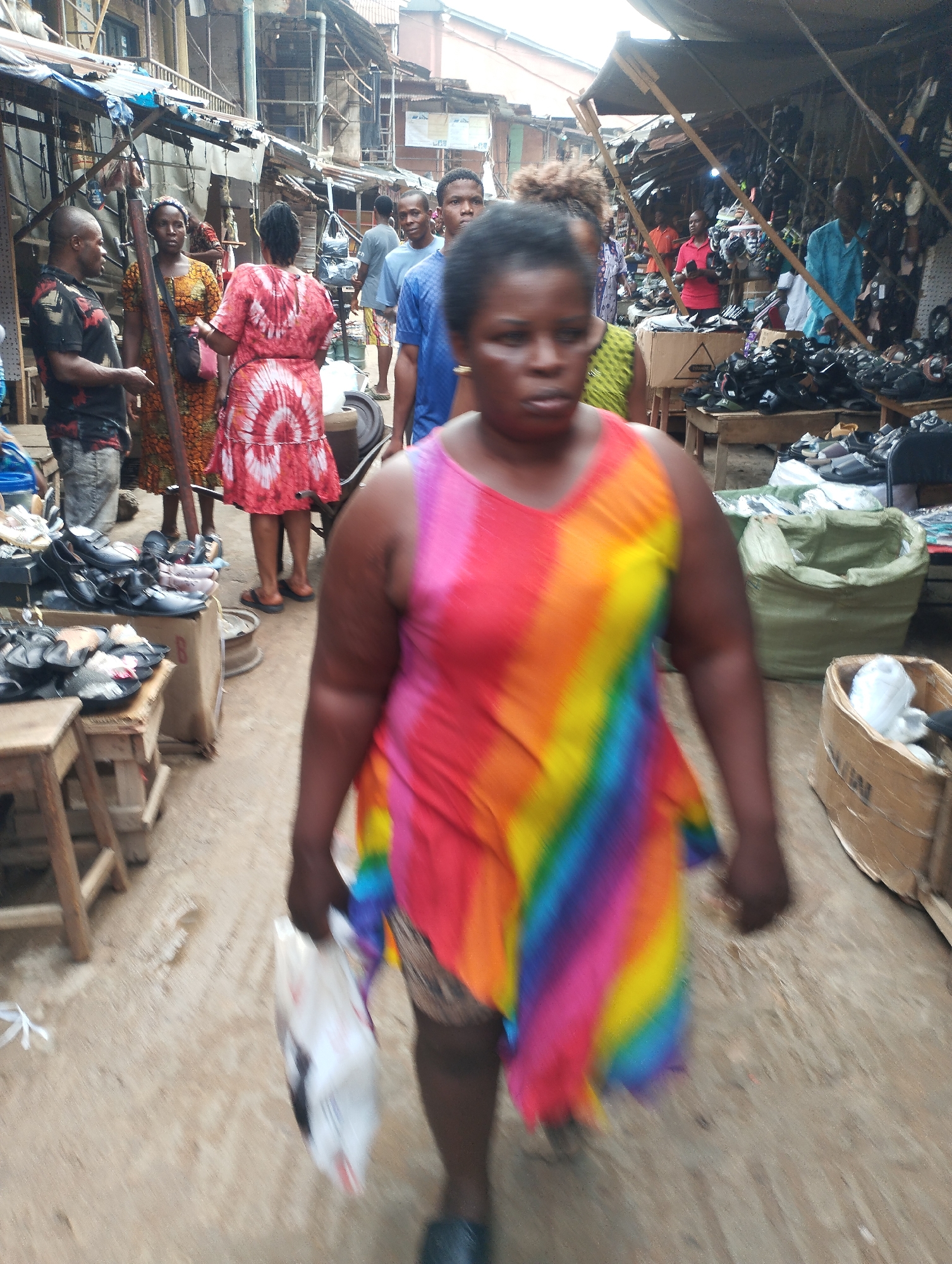 The image depicts a crowded market scene in Fegge, Nigeria. In the foreground, a woman wearing a rainbow-colored dress walks towards the camera, carrying a white plastic bag. Behind her, a dense crowd of people moves in both directions amidst stalls displaying goods, primarily shoes. Buildings constructed with corrugated metal sheets and wooden support structures are visible in the background. The scene is lit by natural light, giving the impression of a busy, open-air marketplace.