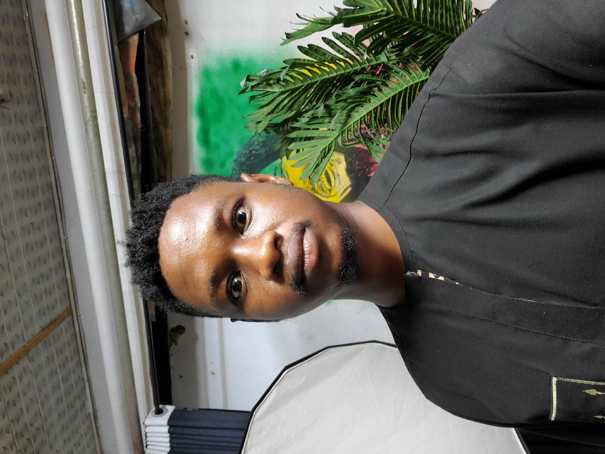 A medium shot depicts a man with brown skin and short, curly black hair. He is wearing a black shirt with gold detailing on the front pocket. He appears to be in an indoor setting. The background is a wall that features green and yellow paint. A potted plant is visible behind the man. A softbox lighting apparatus is also visible in the lower part of the frame. The location is Mararaba, Nigeria.