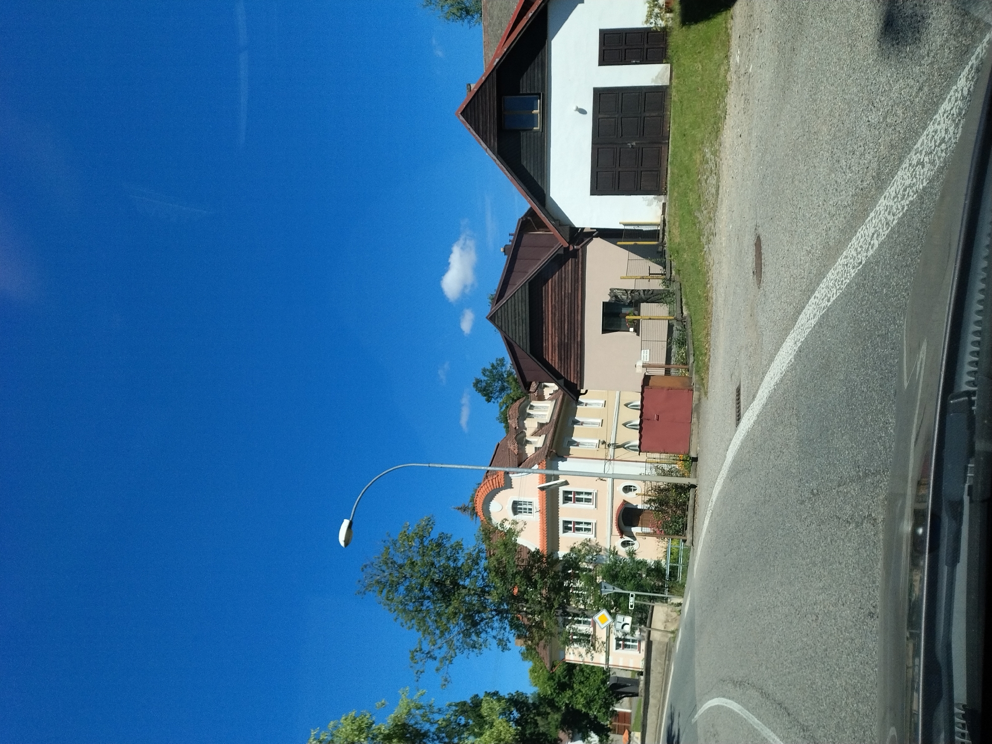 The image shows a street scene in Čkyně, Czechia. On the right side of the road, there are two buildings: one is a white garage with a large brown door and a smaller brown door on the right, and the other is a beige building with a brown roof. In the background, there is a light yellow building with an orange-tiled roof, featuring multiple windows and an arched doorway. A street lamp is visible on the left side of the road, with a clear blue sky above and some green trees and foliage. The road itself is paved and has white lane markings. The foreground shows the dashboard and windshield of a vehicle, indicating the photo was taken from inside a car.