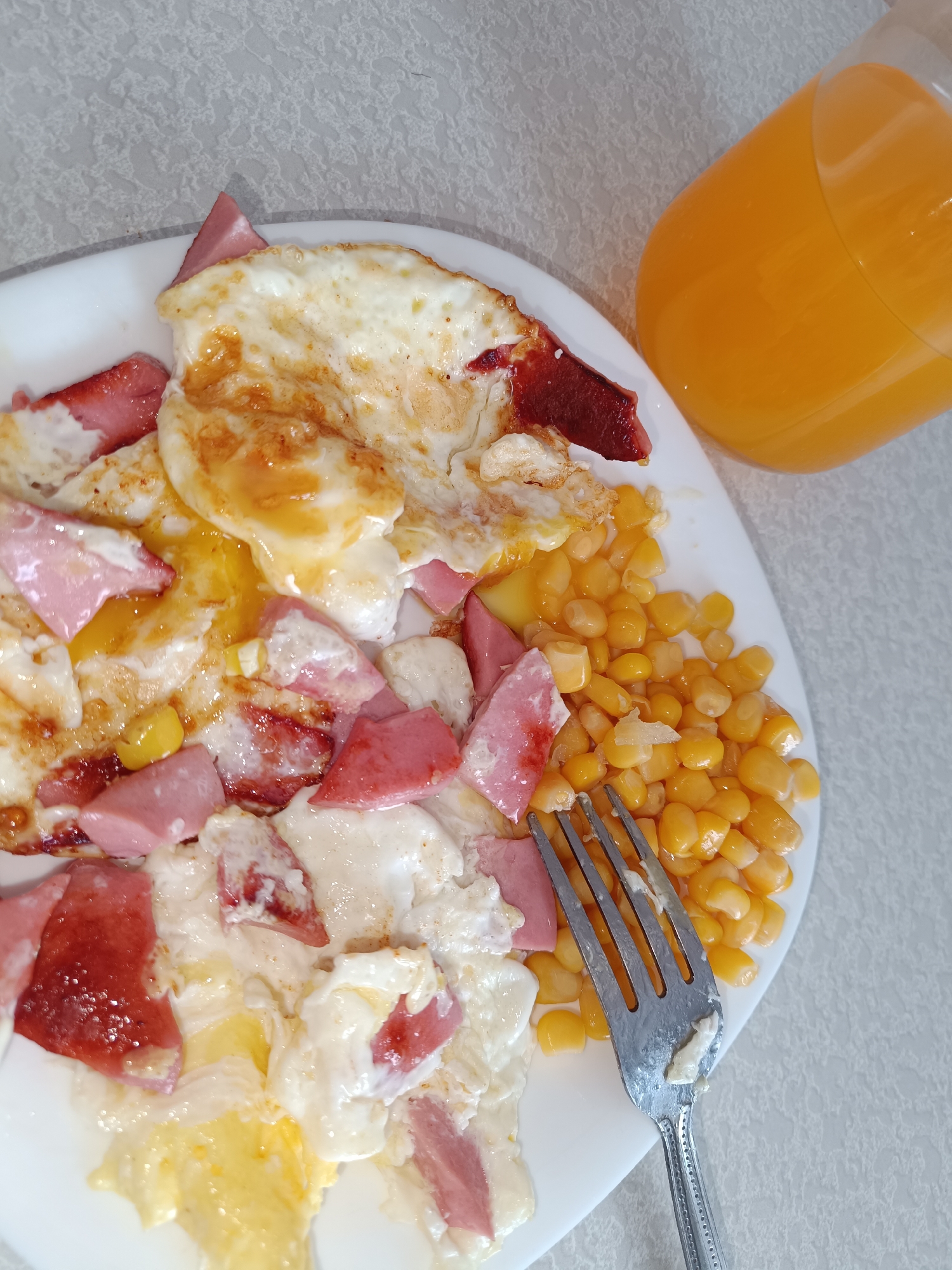 A plate of food, potentially breakfast, is displayed on a white surface. The plate contains fried eggs, diced ham or sausage, and corn. A silver fork is on the plate with some food residue. Next to the plate, there is a large glass of orange juice. The background is a light-colored, textured surface. There is no indication of the location being Tobolsk, Russia. No people are visible.