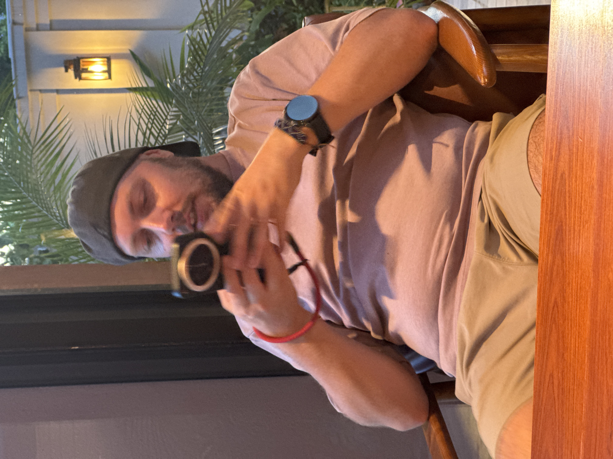 A medium shot captures a man sitting in a wooden chair in what appears to be an outdoor dining setting. He is wearing a light pink t-shirt, beige shorts, and a dark baseball cap. The man is holding a small digital camera in his hands, looking down at it as if to operate it. He has a beard and his skin tone is fair. On his left wrist, he wears a dark watch with a round blue face. On his right wrist, he has a thin red bracelet. The background shows a wall with an outdoor lantern, palm fronds, and what looks like a window or doorway. The scene is lit by the ambient light of the lantern. There is no visible connection to Nam Hoa Lư Ward, Vietnam in the image itself.