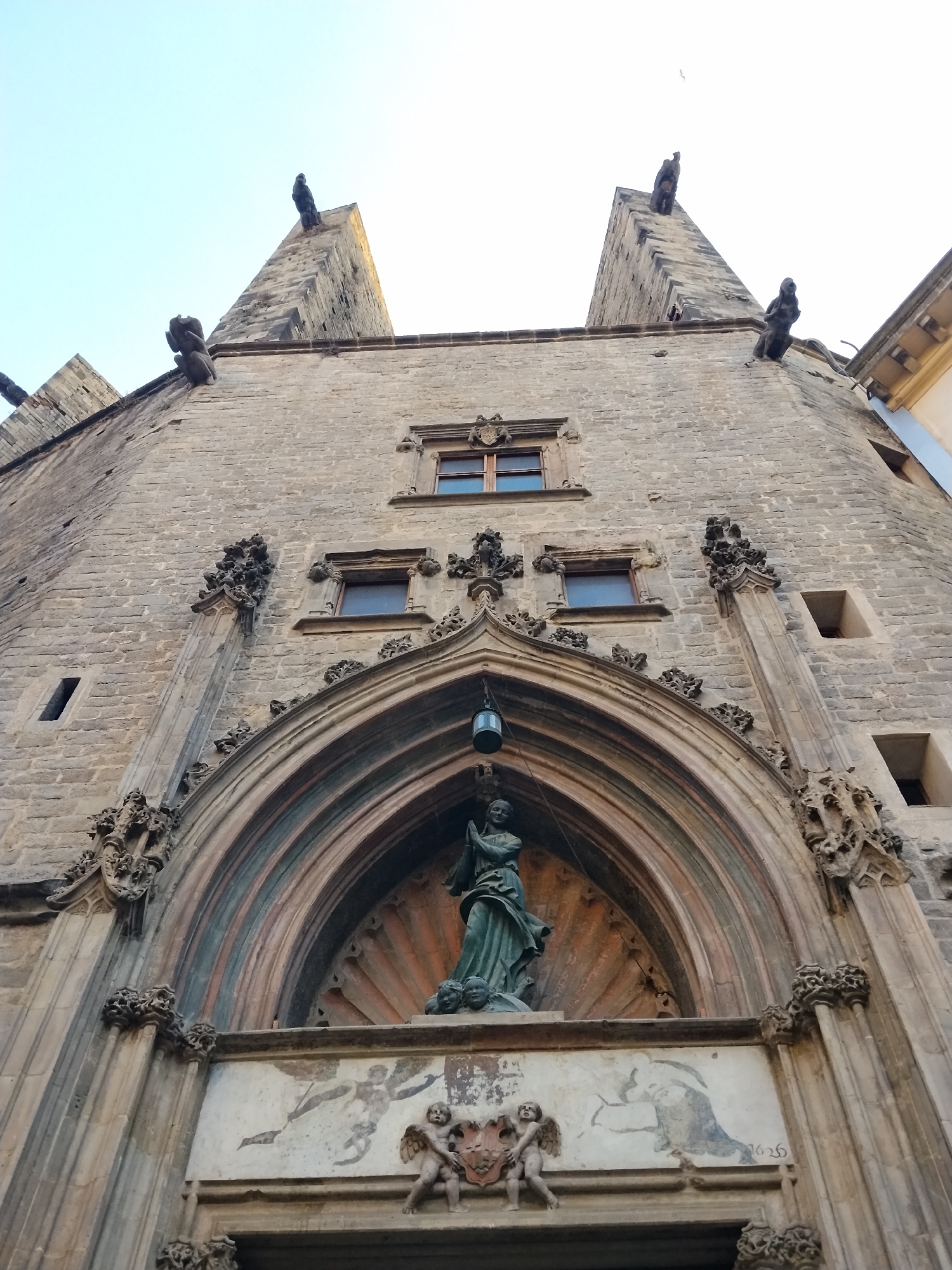 This low-angle photograph captures a majestic, ornate facade of a historical building, likely a church or cathedral, in Barcelona, Spain. The stonework is intricately carved with Gothic architectural elements, including pointed arches, decorative moldings, and grotesque gargoyles perched on the roofline. The building's facade is constructed from weathered, light-colored stone, with darker stone accents forming the arched entryway and window frames.
Dominating the center of the facade is a large, recessed arched portal. Within this archway, a dark green statue of a figure, possibly a religious icon, stands with hands clasped in prayer. Below the statue, a painted frieze depicts winged cherubs and a figure holding a banner, with the year "1626" visible to the right, suggesting the building's age and historical significance. Two arched windows with darker frames are visible above the portal, and above these, two smaller rectangular windows. Two pointed spires, topped with carved figures, rise dramatically from the upper corners of the building, framed against a pale, clear sky. The lighting suggests it is daytime, likely late afternoon or early morning, given the soft, diffused light. The overall impression is one of grandeur, history, and architectural artistry.