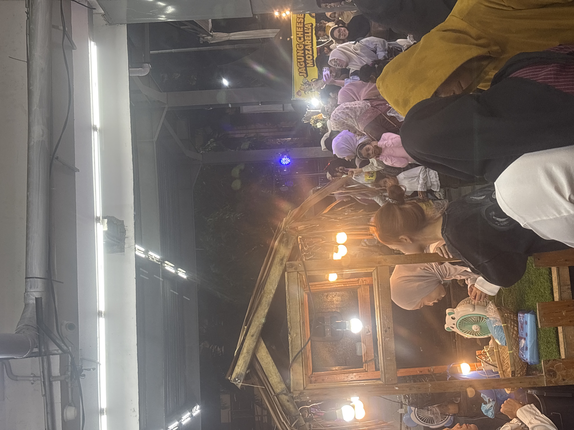 The image depicts a crowded outdoor night market or event in Lhokseumawe, Indonesia. Overhead, exposed grey pipes, electrical conduits, and cables are visible against a light-colored ceiling, with several long, bright fluorescent light fixtures illuminating the area. A small, dark security camera is mounted on the ceiling, and a section of damaged structure is visible on the upper right.
In the mid-left, a rustic wooden stall with a simple roof is prominently featured, illuminated by multiple bare incandescent light bulbs. Inside the stall, a white, animal-shaped fan with a teal blade rests on a green, artificial grass-like surface, alongside a smaller black fan, woven baskets, and blue-wrapped packages. A woman in a light grey hijab is visible behind the counter, looking towards the right.
The midground and right side of the image are densely populated with people, primarily women wearing various colored hijabs (including pink, grey, light blue, and white) and children. A young girl in a pink top is centrally positioned, looking towards the viewer. In the lower right foreground, the backs of two individuals are visible, one in a black head covering/hoodie and another in a yellow hoodie. On the far right, a yellow banner with red text advertises "JAGUNG CHEESE MOZARELLA" (Corn Cheese Mozzarella) and displays a corn graphic. A blue, star-shaped light fixture is visible in the background, to the left of the banner. The crowd is standing, observing the surroundings, and engaging with the market environment. The scene is illuminated by numerous artificial light sources, indicating a nighttime setting with dark foliage visible in the distant background.