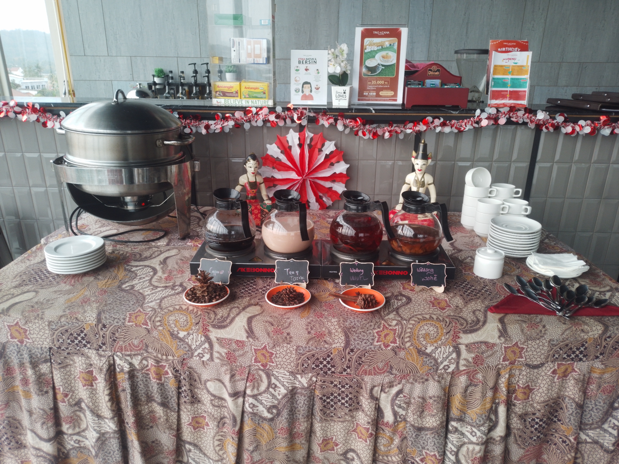 The image shows a buffet table at a cafe or restaurant, likely in Bumirejo, Indonesia, set up for a beverage service. The table is covered with a batik-patterned tablecloth. Prominently displayed are four glass carafes, each with a black lid, placed on individual heating elements. The carafes contain different beverages: the first appears to be coffee, the second a creamy liquid like milk tea or a latte, the third a dark red liquid like tea or herbal infusion, and the fourth a similar dark red liquid. Small orange dishes with pinecones and black chalkboards with handwritten labels in Indonesian, indicating "Coffee", "Tea & Tarik", "Wedang Uwuh", and "Wedang Jahe", are placed in front of each carafe.
To the left of the carafes is a large, silver multi-tiered food steamer. To the right, a stack of white ceramic cups and saucers is visible, along with a white sugar dispenser. A bundle of spoons is arranged on a red napkin.
In the background, a grey tiled wall serves as the backdrop. Decorative elements include a red and white paper fan and two traditional Indonesian figurines. Various products and signs are displayed on shelves behind the buffet setup, including boxes of "Antangin Dolpin", a health product, and promotional materials for "Trio Azana". A window on the left offers a view of greenery and distant buildings. The overall scene suggests a self-service beverage station offering hot drinks.