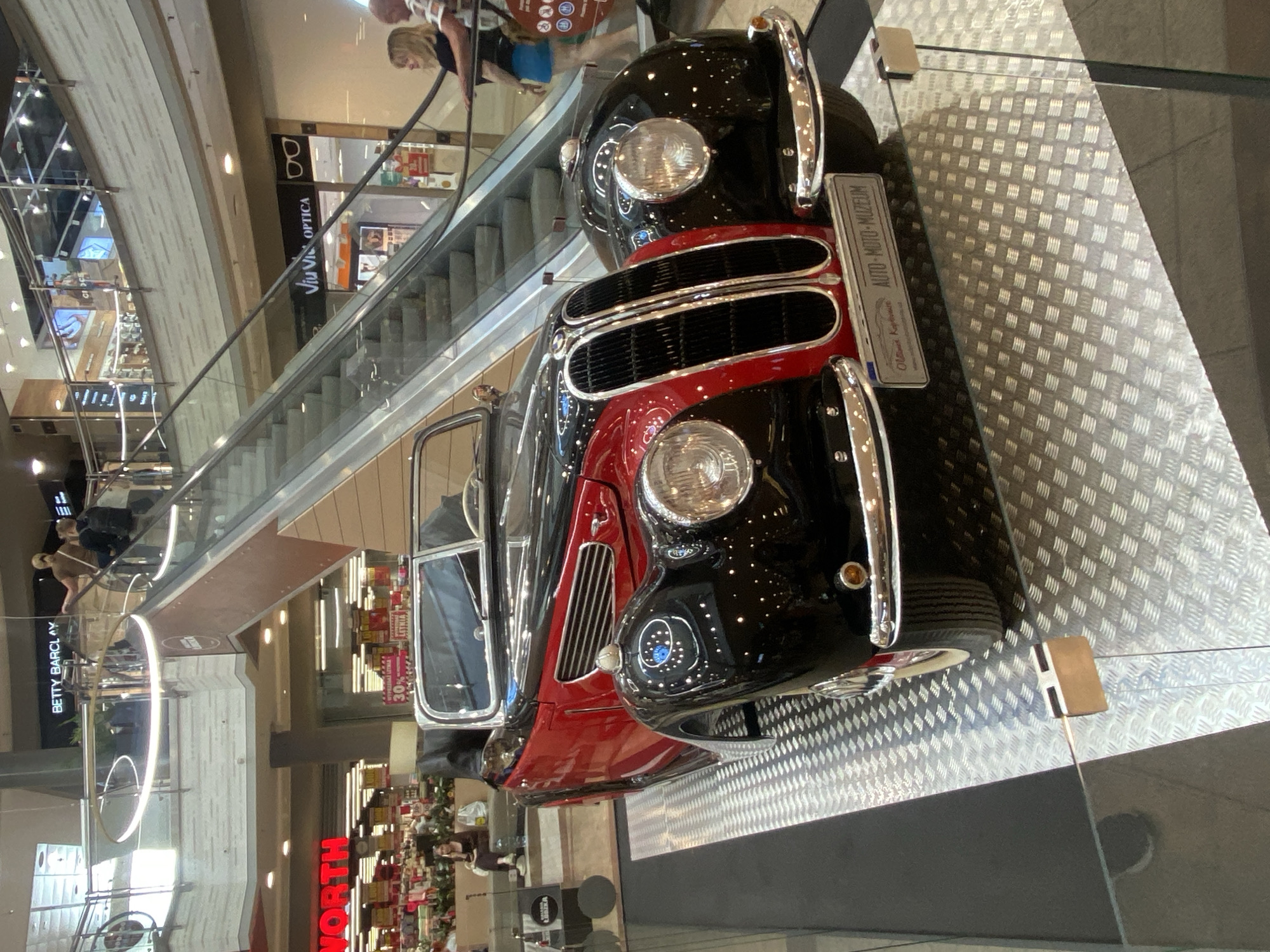 This is a vertical, medium-shot photograph taken indoors, likely in a shopping mall or exhibition space in Łódź, Poland, as indicated by the text "LODZKA" and the mall-like architecture. The primary subject is a vintage two-tone black and red car, possibly a convertible due to its open top. The car is prominently displayed on a metallic checkered platform. A sign attached to the car reads "AUTO-MOTO-MUZEUM" and "Oltimet Kaptive," suggesting it is part of a museum exhibit.
In the background, an escalator is visible, with several people casually walking or standing on it. One woman with blonde hair is looking towards the car, while another person is partially visible behind her. Below the escalator, further into the mall, shops are visible, including one with a sign that says "BETTY BARCLAY." Another sign displays "NORTH" in large red letters, possibly indicating a department store or the name of the mall. A red sign with white text also shows "30%," indicating a sale. The overall atmosphere appears to be casual and commercial, with no strong indicators of time of day or weather. The lighting is artificial, typical of an indoor shopping environment.