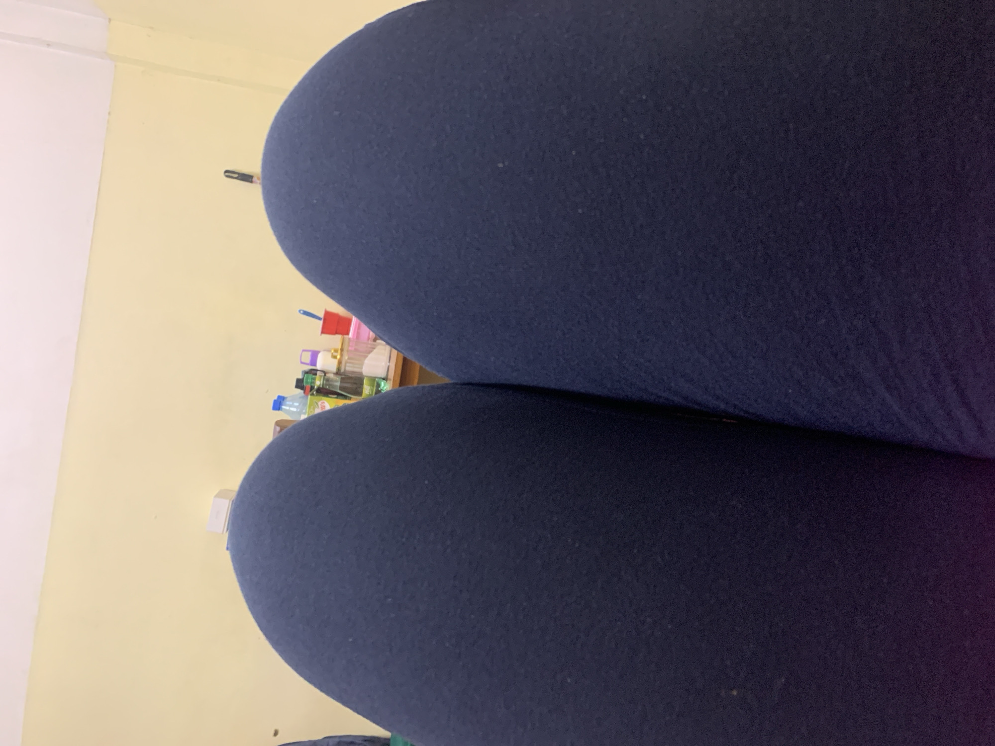The image shows a close-up view of two legs covered in dark blue leggings. In the background, a yellow wall is visible, along with a shelf containing various bottles and containers. The setting appears to be indoors, possibly in a home or apartment. There are no clear indicators to definitively place the image in Dar es-Salaam, Tanzania.