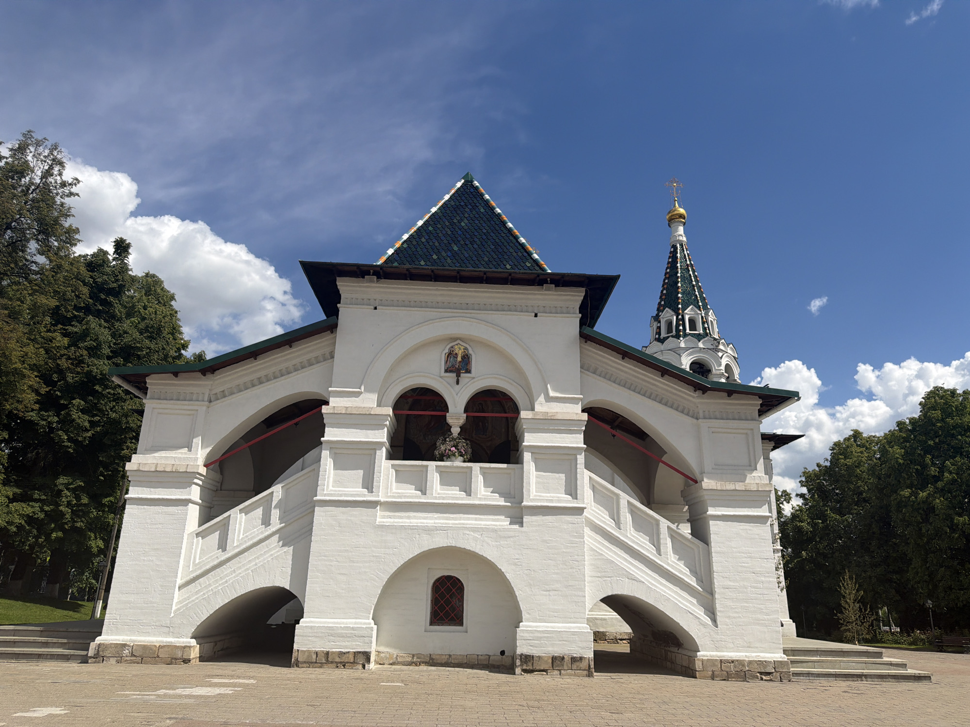 The image displays a prominent white architectural structure under a clear blue sky with scattered white clouds, characteristic of a sunny day. This building features a grand, double-flight staircase leading to an elevated, open-air gallery. The staircase, constructed of white brick or stucco, has arched passageways underneath and is flanked by solid balustrades.
The upper gallery is supported by thick pillars and defined by a series of arches, with red railings or trim visible within the arched openings. Above the central arch of the gallery, a small, colorful religious icon or fresco is inset. On the ground level, beneath the gallery, there is a small arched window with a red-gridded frame. The building's roof is steep and pyramidal, covered with patterned dark green and lighter green or blue tiles.
To the right and slightly behind this structure, a tall, slender bell tower rises, also white, with a similarly patterned, conical spire. The bell tower is topped with a gleaming golden onion dome and a cross, reflecting the sunlight. Lush green trees frame both sides of the scene, suggesting a park-like or landscaped setting. The foreground is paved with light-colored bricks or cobblestones. The architectural style, including the golden dome and cross, indicates a Russian Orthodox religious complex in the Pavlovskaya Sloboda, Russia context. No people or discernible text are visible in the image.
