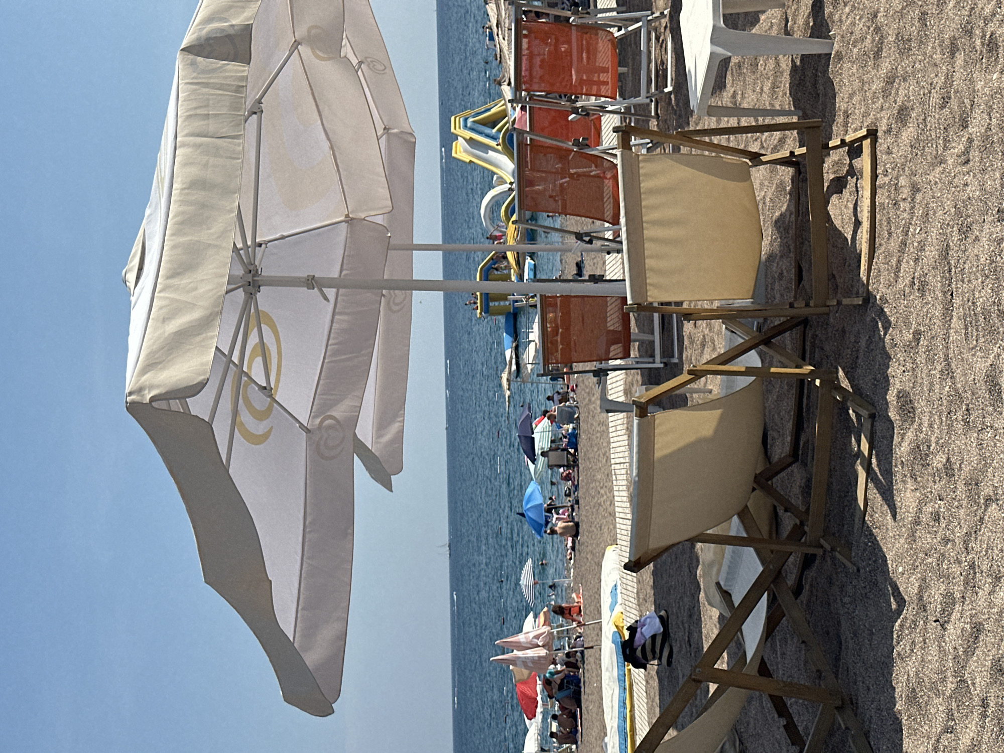 The image depicts a sunny day at a beach in Boreti, Montenegro. In the foreground, several beach chairs are set up on the sand, some of which are occupied by people. A large white umbrella provides shade over some of the seating area. In the middle ground, there is a beach with many people sunbathing and swimming in the ocean. Colorful umbrellas and beach towels are scattered across the sand. In the background, the blue ocean stretches out to the horizon, with a few boats visible. There is also a floating water park with slides. The overall atmosphere is one of leisure and recreation.