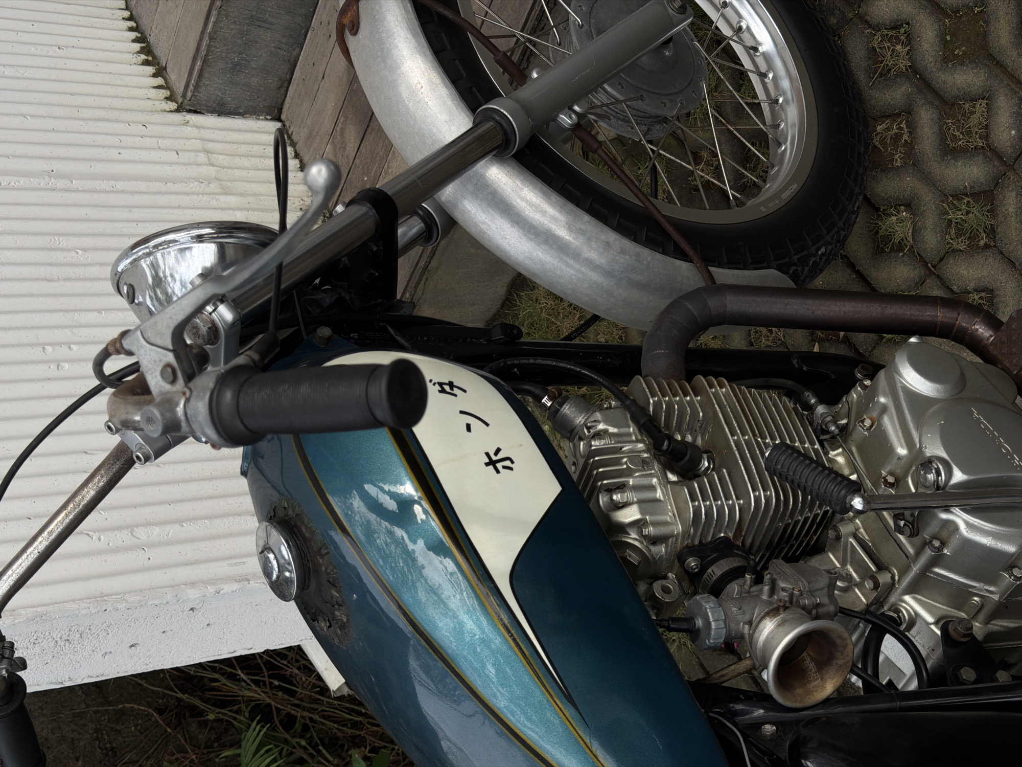 In Mengwi, Indonesia, a close-up, eye-level view captures the right side of a vintage blue and white motorcycle. The motorcycle features a metallic teal gas tank with a yellow and black pinstripe detail, and white lettering that reads "ホンダ" (Honda) in black. A chrome fuel cap is visible on the tank, showing signs of rust and peeling paint underneath. The motorcycle's engine block is silver and appears detailed with fins. To the right, a silver fender covers the front wheel, which has spokes and a black tire. The motorcycle is parked outdoors, with a white corrugated wall in the background and wooden planks and patterned concrete pavers on the ground. Sparse grass grows between the pavers. The lighting suggests it is daytime.