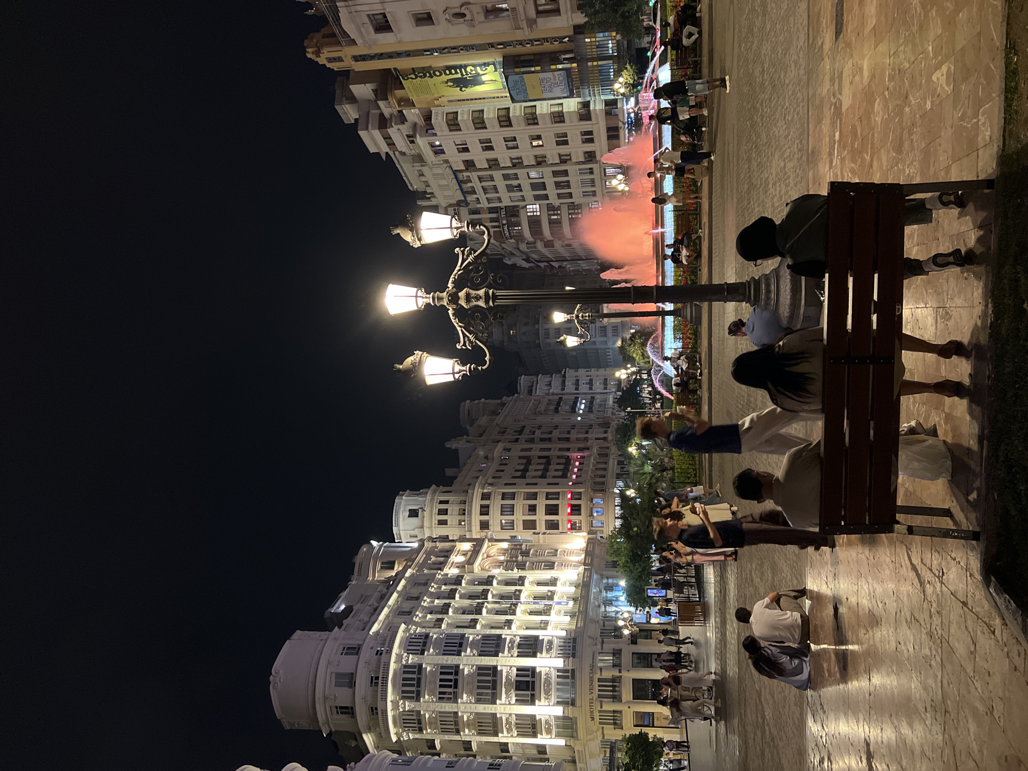 The image shows a nighttime scene in València, Spain, likely in or near the Plaza del Ayuntamiento. Ornate buildings line the left side of the frame, illuminated by lights. A central lamppost stands prominently, casting light on the scene. To the right is a pedestrian walkway or plaza area where people are walking and seated. A bench is occupied by several individuals, some sitting and some standing behind it. In the background, a fountain feature is illuminated with colored lights, creating a pinkish glow in the air. The overall atmosphere suggests a vibrant urban setting during the evening.