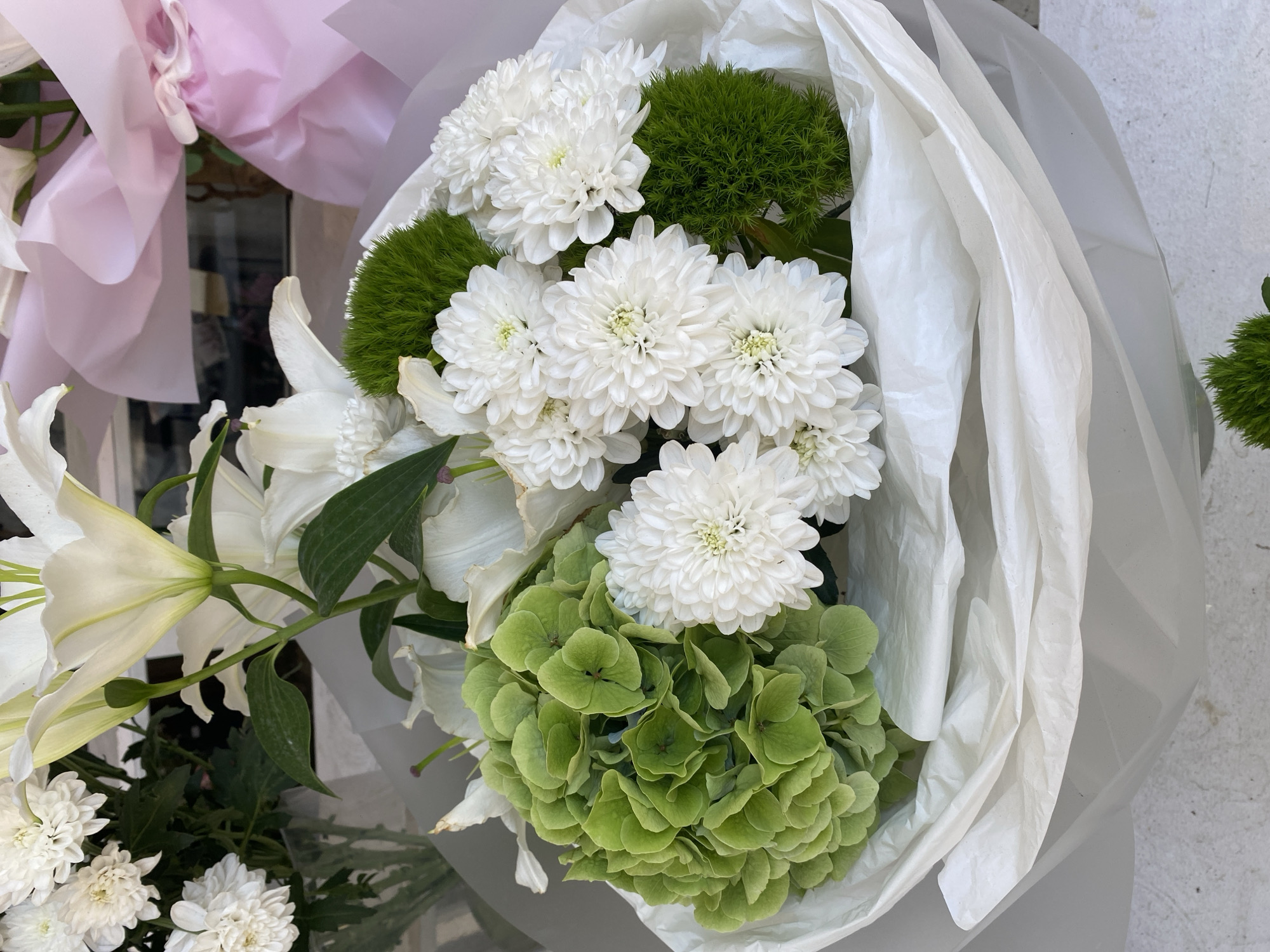The image shows a bouquet of white lilies, white chrysanthemums, green hydrangeas, and green dianthus flowers, arranged in a white paper wrap. The bouquet is placed on a light-colored surface, possibly a table or countertop. In the background, there are glimpses of other flowers and pink tissue paper, suggesting a florist's shop or a gift arrangement. There are no people or specific city landmarks visible in the image.