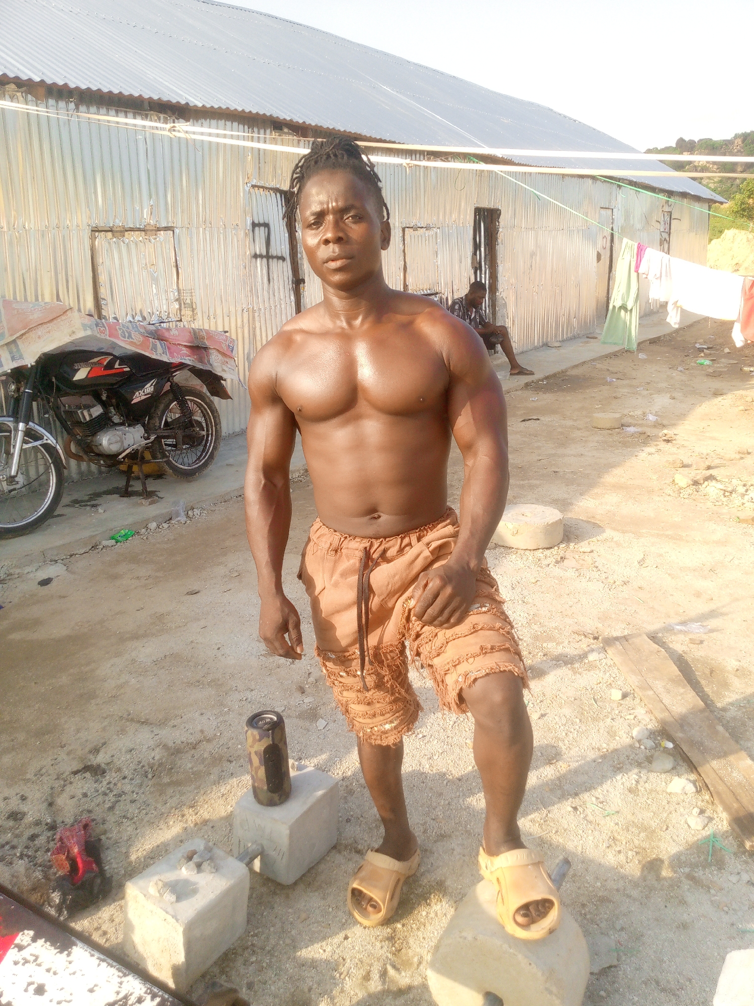 A shirtless man with dreadlocks and muscular physique stands in front of a corrugated metal building in Indo, Nigeria. He is wearing distressed tan shorts and tan flip-flops, with his right foot resting on a concrete block. A small portable speaker with a camouflage pattern sits on another concrete block nearby. To his left, a motorcycle with a red and black design is parked. In the background, another person is seated near the entrance of the building, and laundry hangs on a clothesline. The ground is dirt and scattered with debris.