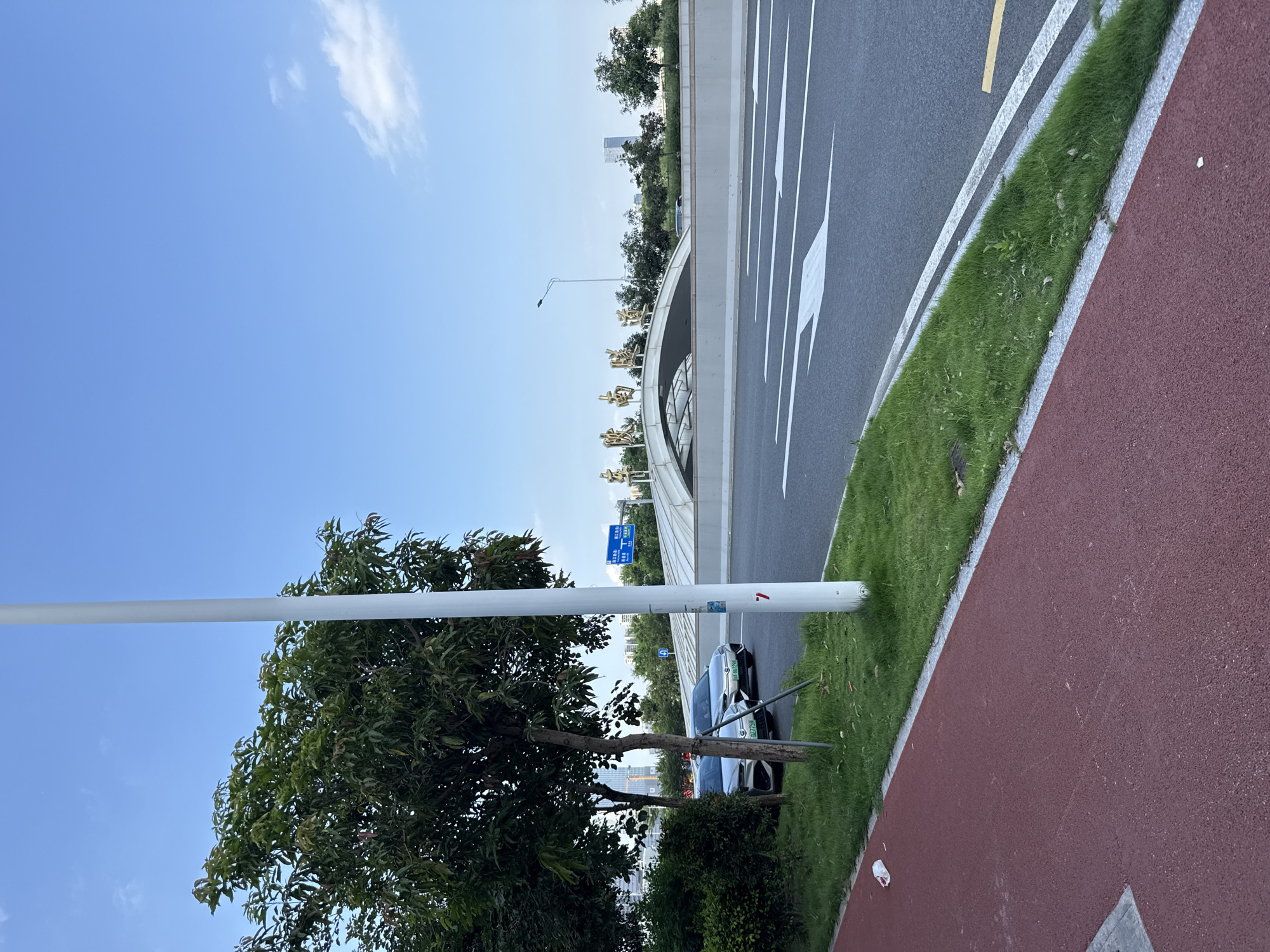 In Haizhu District, China, a clear blue sky with scattered clouds overlooks a modern urban scene. A paved road with white directional arrows dominates the foreground, bordered by a red jogging path and a strip of green grass. Two silver sedans are parked on the grass to the left, partially obscured by a streetlamp. In the background, a white, arching structure spans across the road, adorned with golden Chinese characters. Beyond the structure, trees and modern buildings rise against the sky. A blue traffic sign with white lettering is visible on a pole. The overall impression is one of a well-maintained urban environment on a bright, clear day.