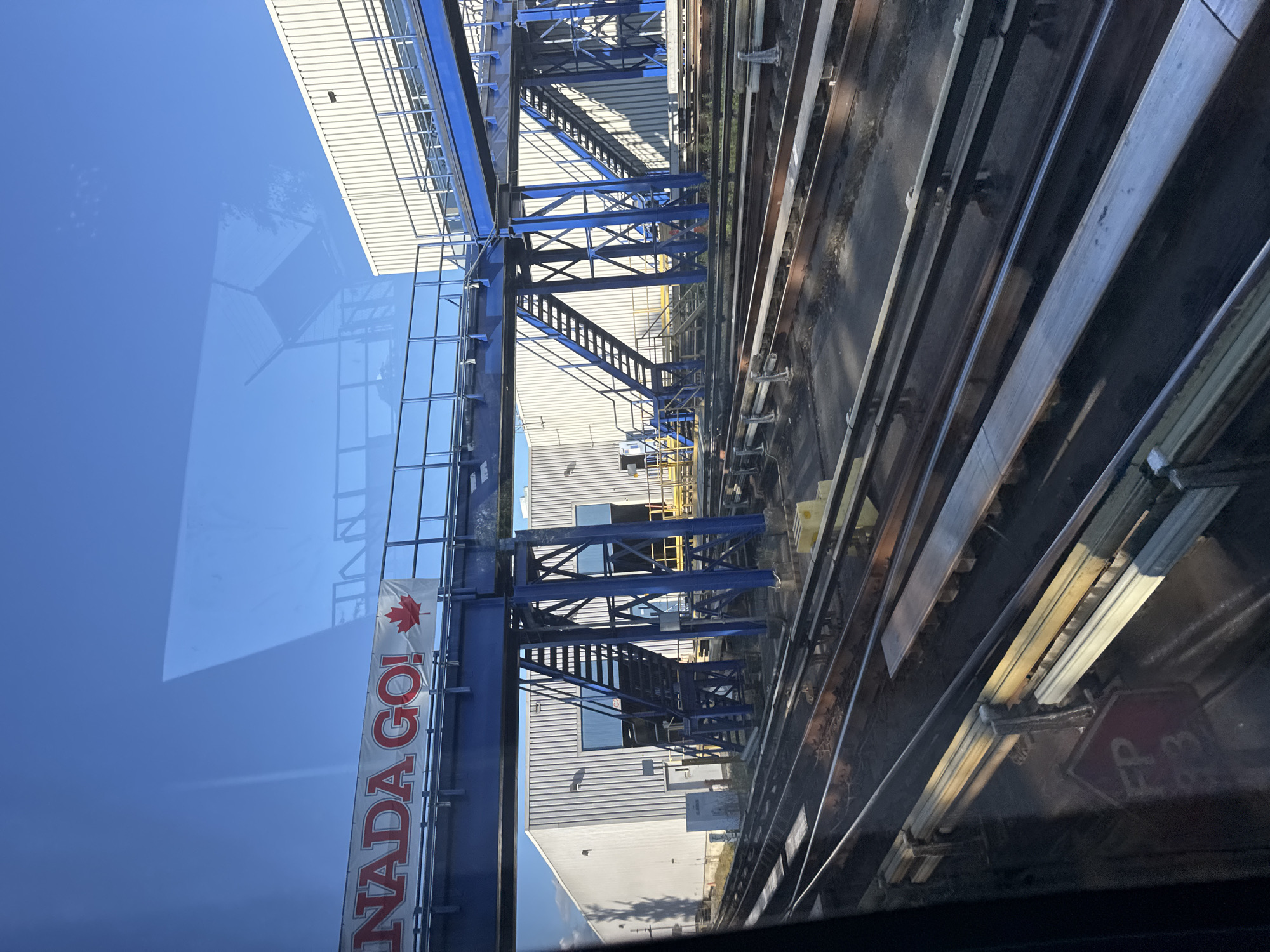 The image, taken from inside a vehicle, captures a view of an industrial area with multiple railway tracks in the foreground. In the mid-ground, a large, white industrial building with corrugated siding is visible. Adjacent to it, a complex structure with blue painted metal supports and stairs leads up to a walkway or bridge. A banner with red lettering that reads "CANADA GO!" and a Canadian flag is hung on the walkway. The sky above is clear and blue, with some light reflections on the window. The scene is likely located in Burnaby, Canada, given the banner. There are no people visible in the image.