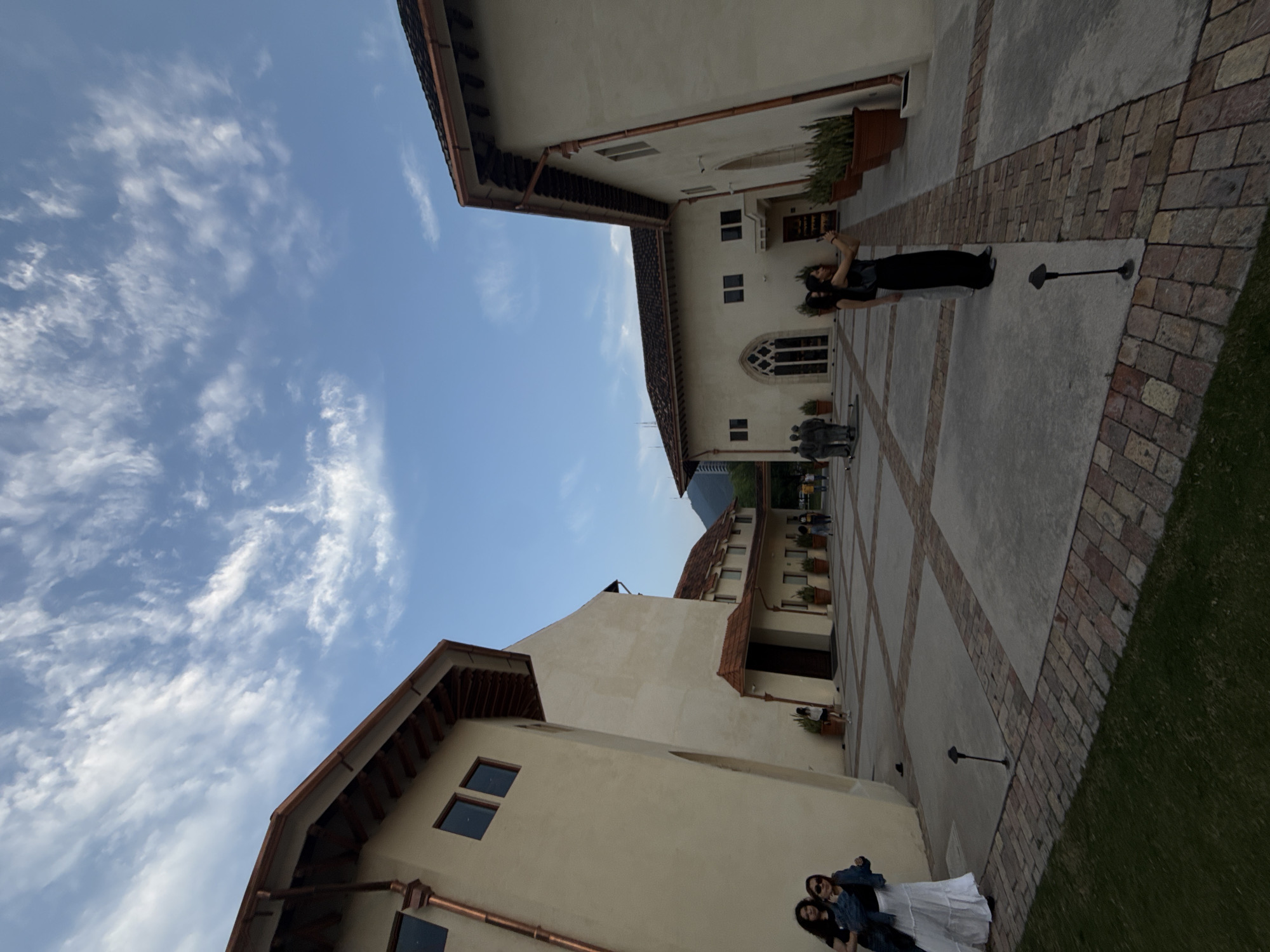 The image shows an outdoor courtyard area with several people present, located in San Pedro Garza García, Mexico. The architecture of the buildings surrounding the courtyard is European-inspired, with beige walls, copper rain gutters, and red tile roofs. In the lower part of the image, two women are standing together, dressed in dark shirts and light-colored skirts. Near the center of the image, further up the courtyard, another woman is standing, holding her phone up as if taking a picture. There are brick pathways that intersect with concrete walkways. At the end of the courtyard, closer to the buildings, a group of people is standing near a statue. The sky is blue with wispy clouds.