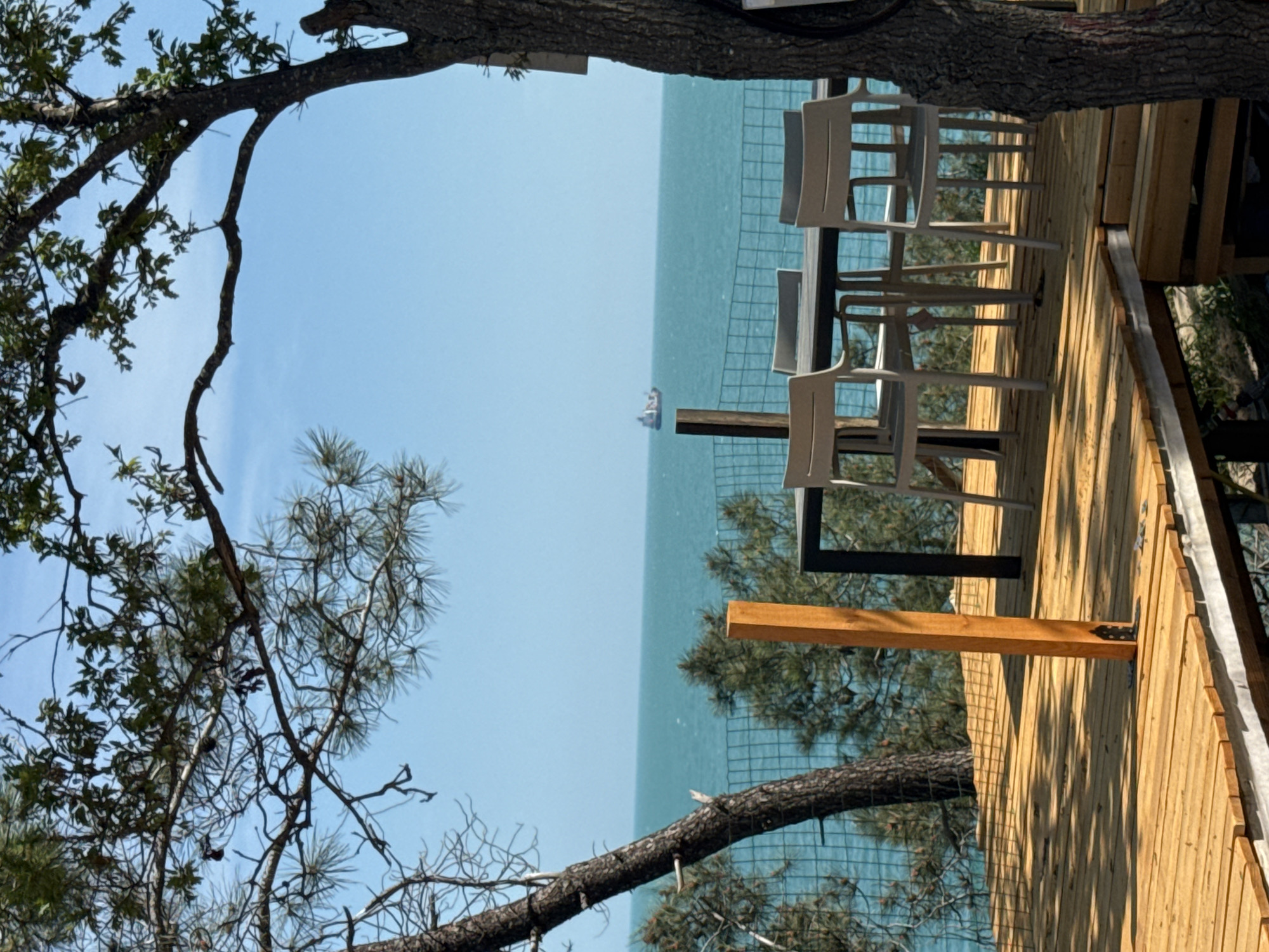 View from a wooden deck overlooking the sea in Varna, Bulgaria, framed by trees. The deck features a table with four chairs. A ship is visible in the distance on the water. A wire fence runs along the edge of the deck.